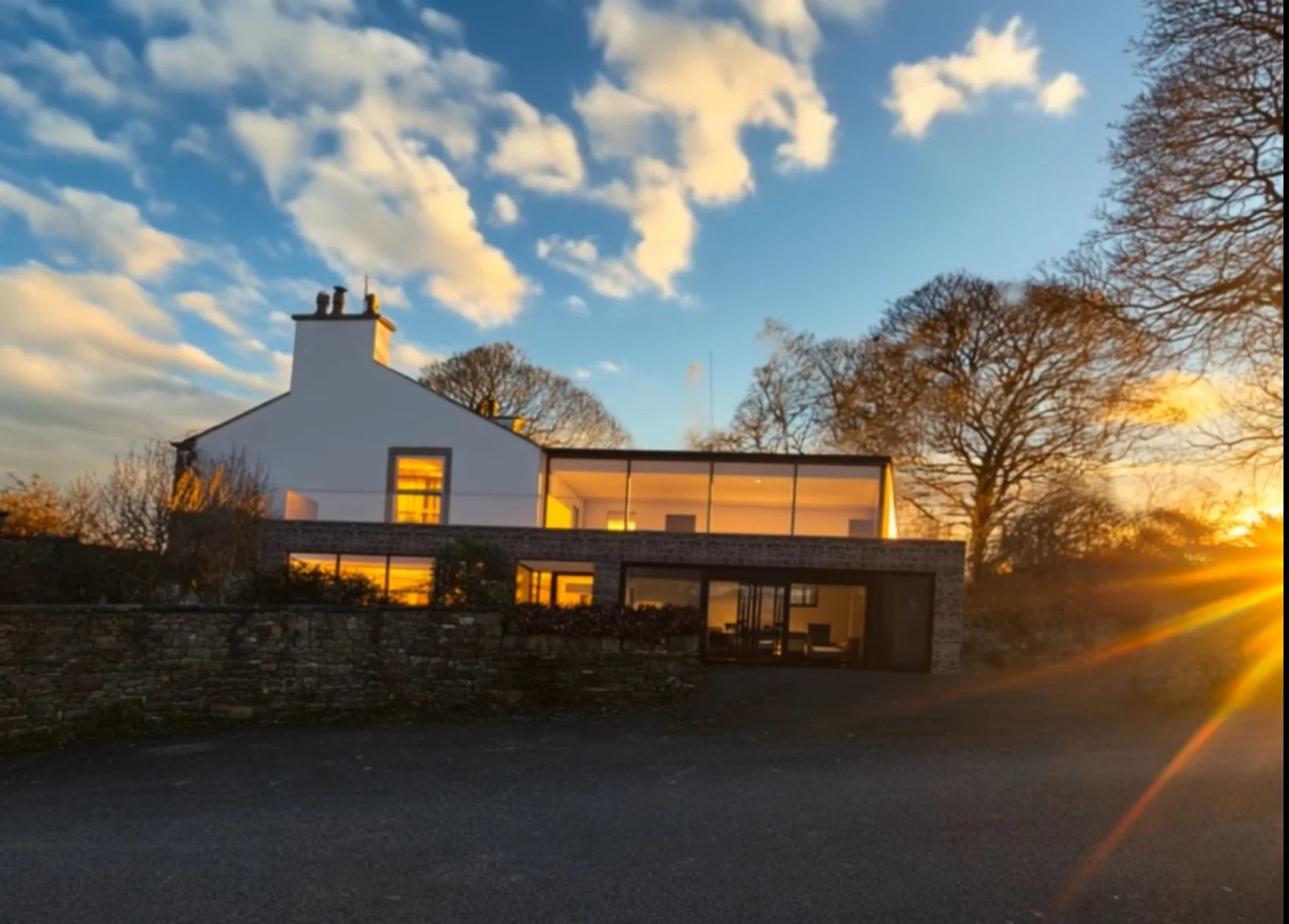 A modern house with large glass windows and a traditional white house in the background, set against a sunset with trees and a partly cloudy sky