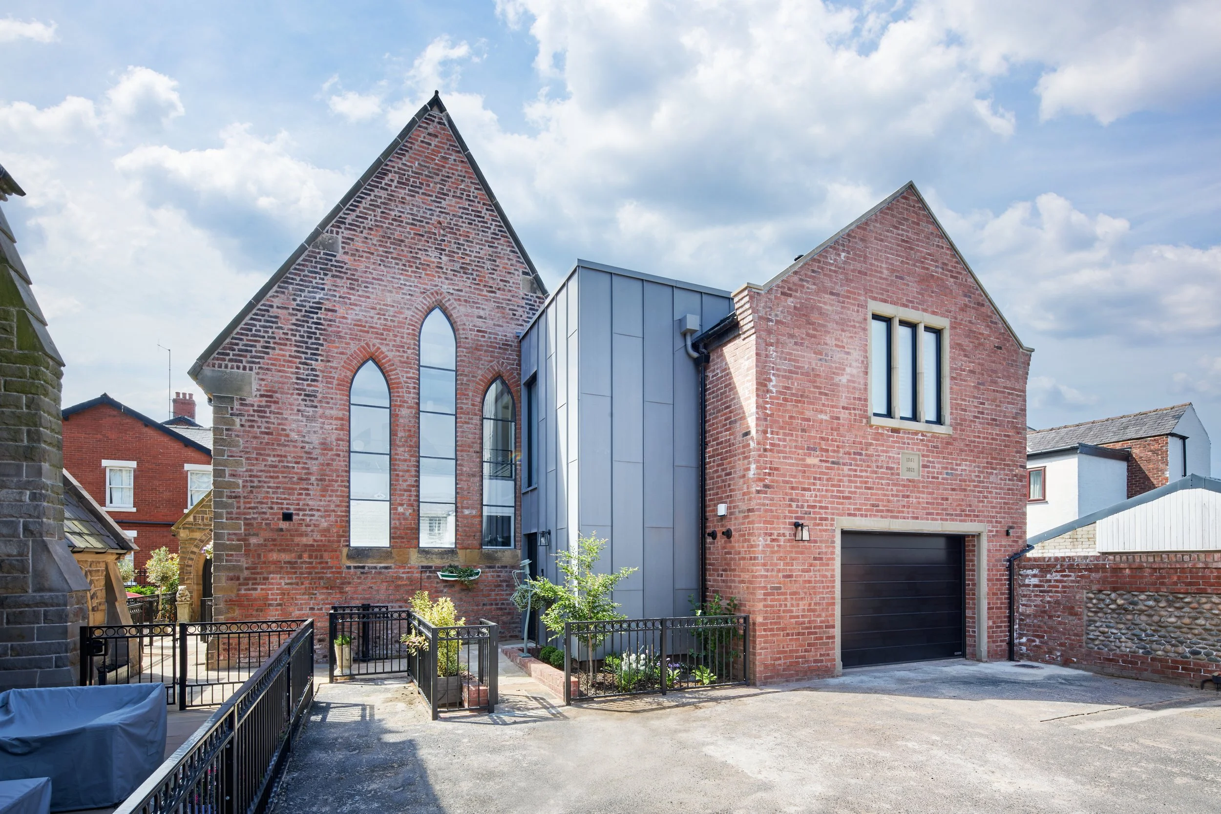 A modern brick house with large arched and rectangular windows, metal panels on the side, a black garage door, and a small front garden with plants and a fence under a partly cloudy sky.