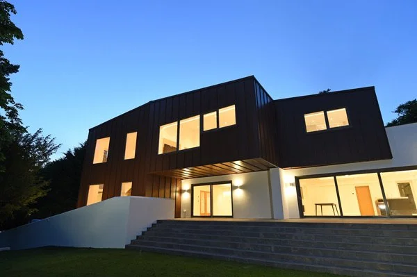 Modern two-story house with black and white exterior, large windows, illuminated porch, and steps leading to entrance, situated in a landscaped yard during twilight.