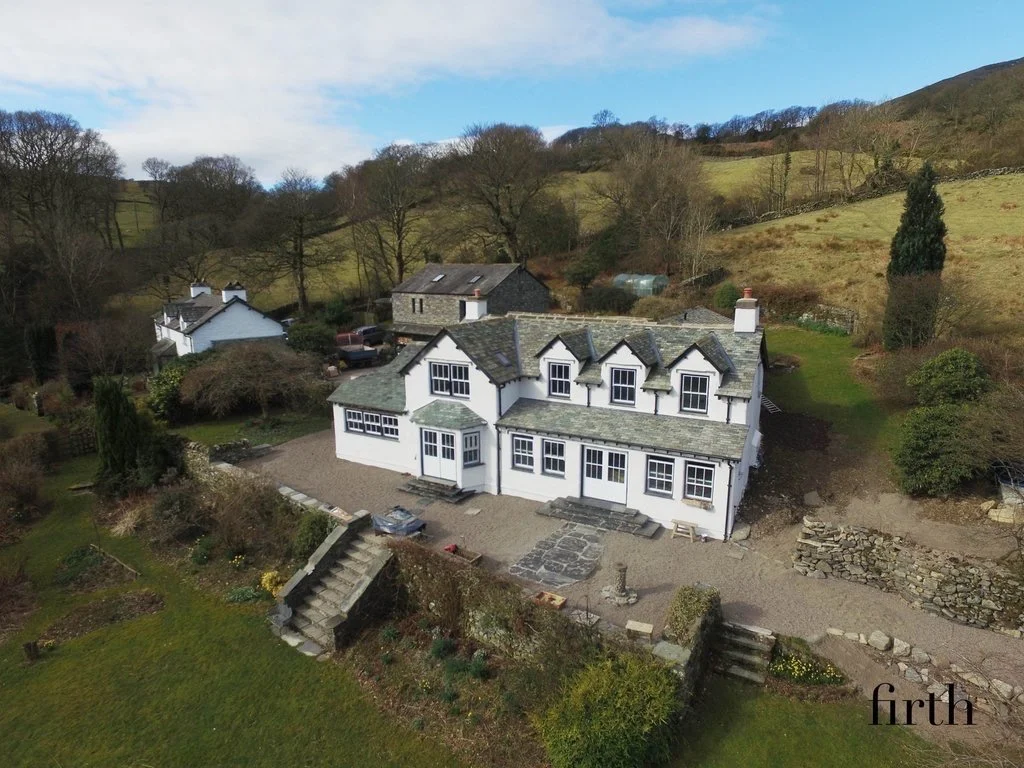 A large white house with black roof shingles, multiple chimneys, and numerous windows, situated on a sloped landscape with surrounding trees, bushes, and a gravel yard.