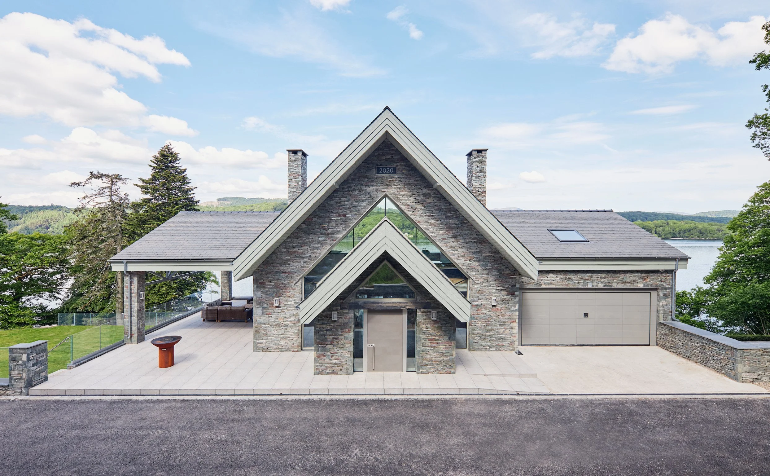 Modern house with stone facade and steeply pitched roof, overlooking a lake with greenery in the background, under a partly cloudy sky.