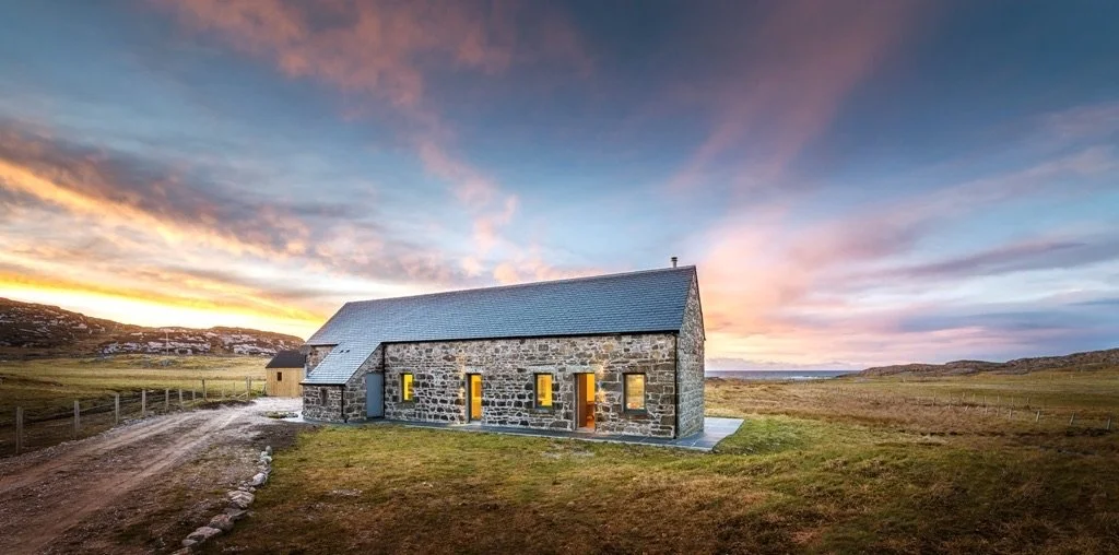 A stone house with a pitched roof situated in a rural landscape at sunset.