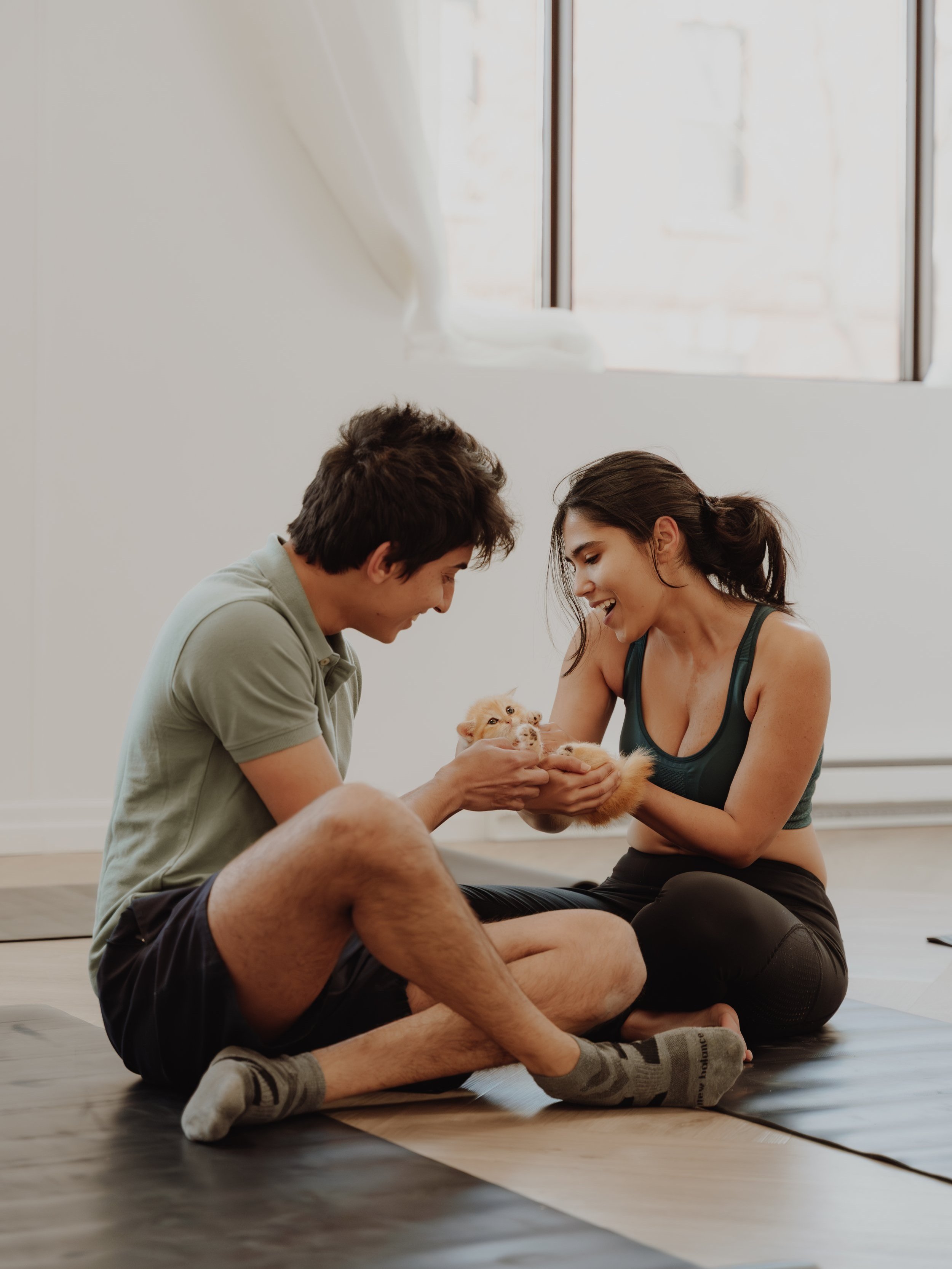 A man and woman sitting on the floor in a bright room, playing with an orange kitten. The woman is holding the kitten while the man reaches out to pet it. Both are smiling and appear happy.