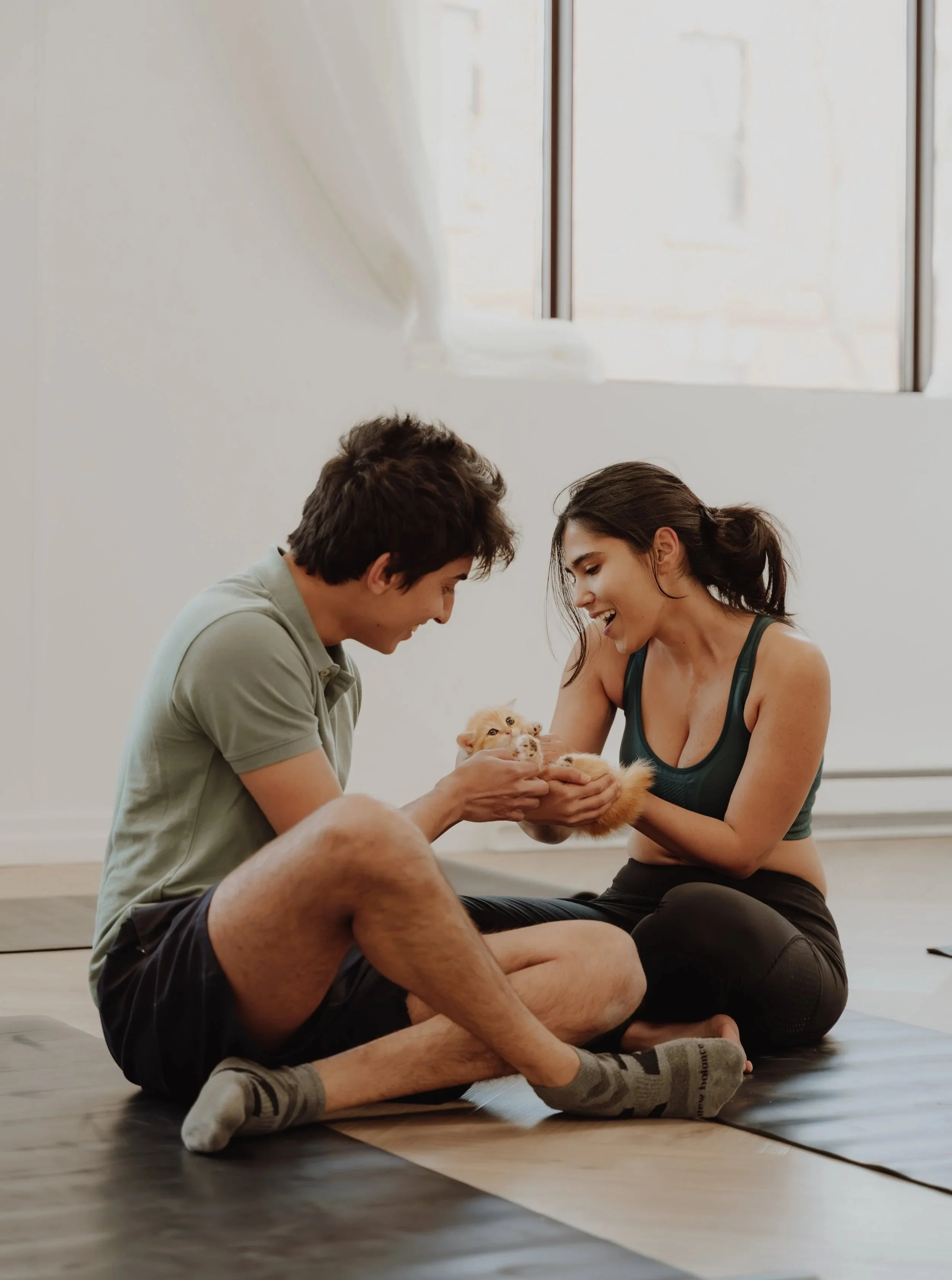 A young man and woman are sitting on the floor in a bright room, playing with a tiny fluffy puppy together.