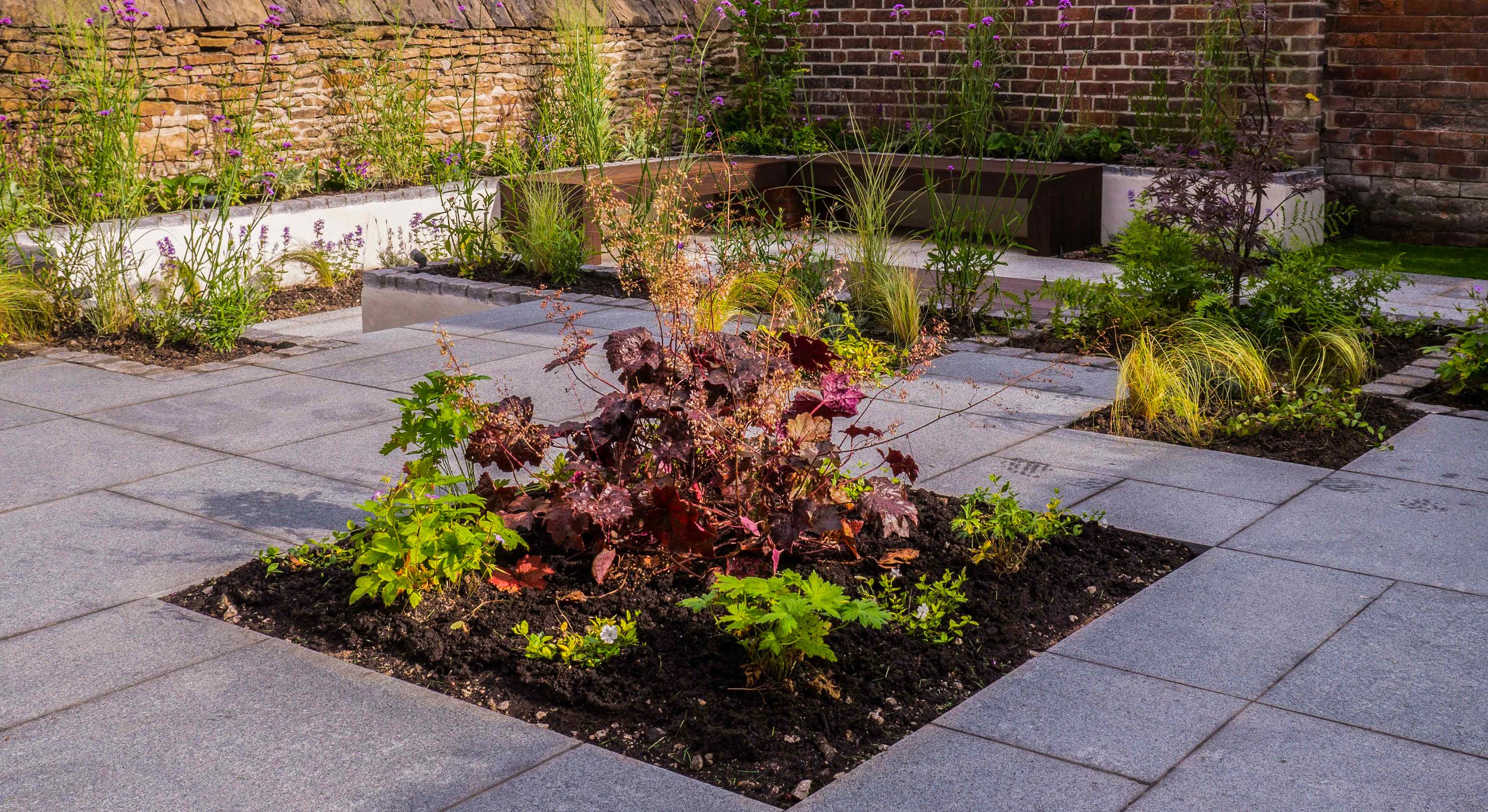 Brown, wooden bench in the Chic garden surrounded by soft planting.  Different levels and light grey granite paving
