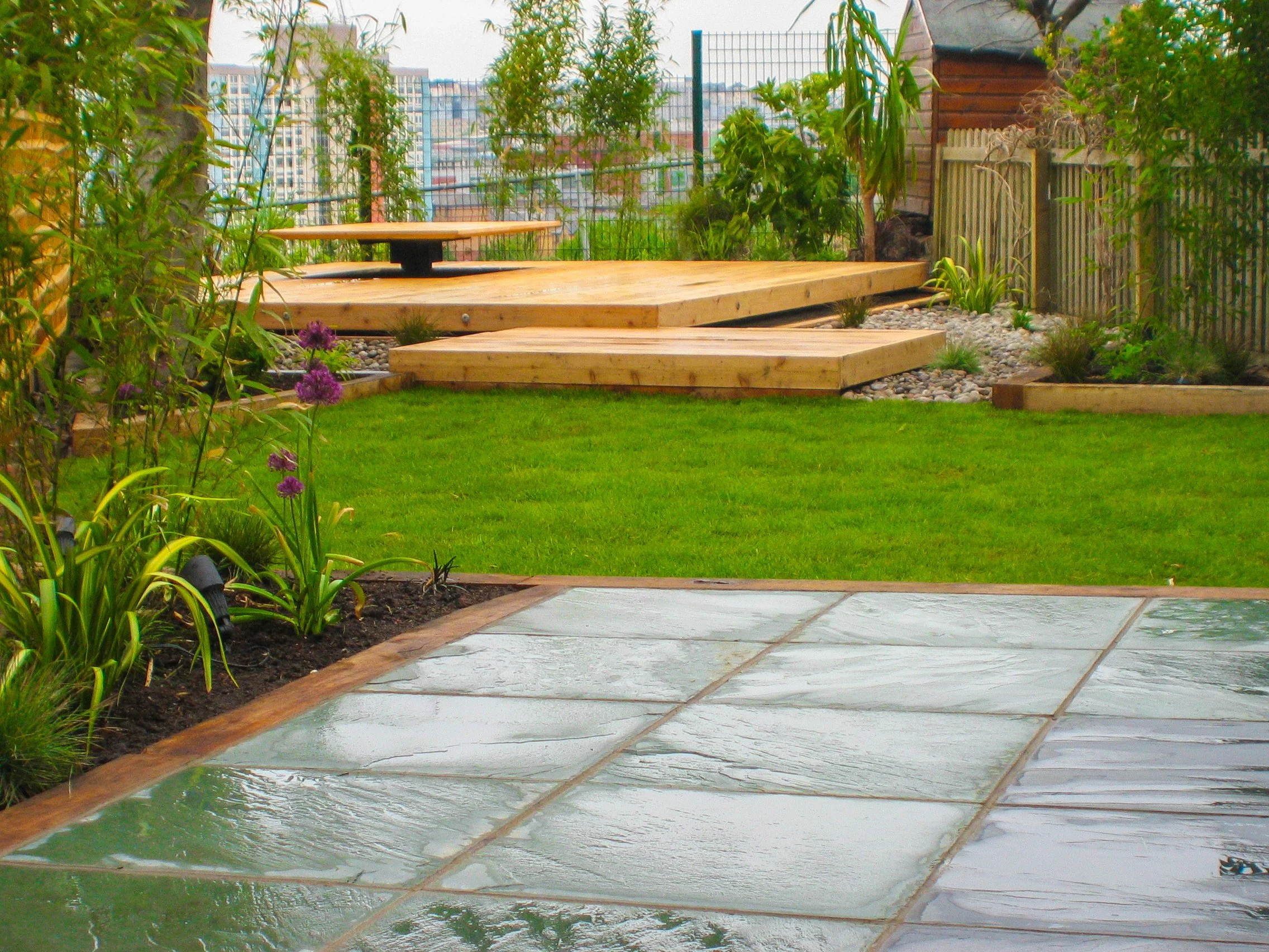 Image of a garden with a lush green lawn and paving in the foreground.   light timber raised decking in the background.