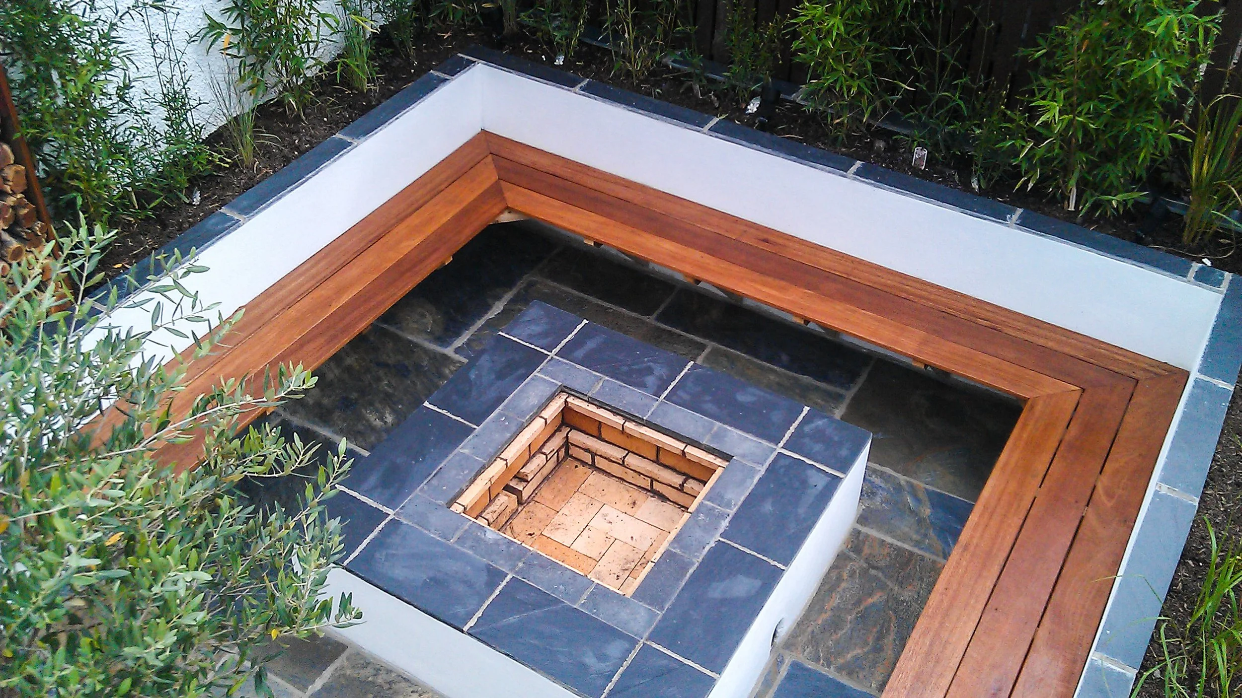 An outdoor fire pit with a square brick interior, surrounded by dark blue and black tiles, with a wooden bench on one side and plants along the edge.