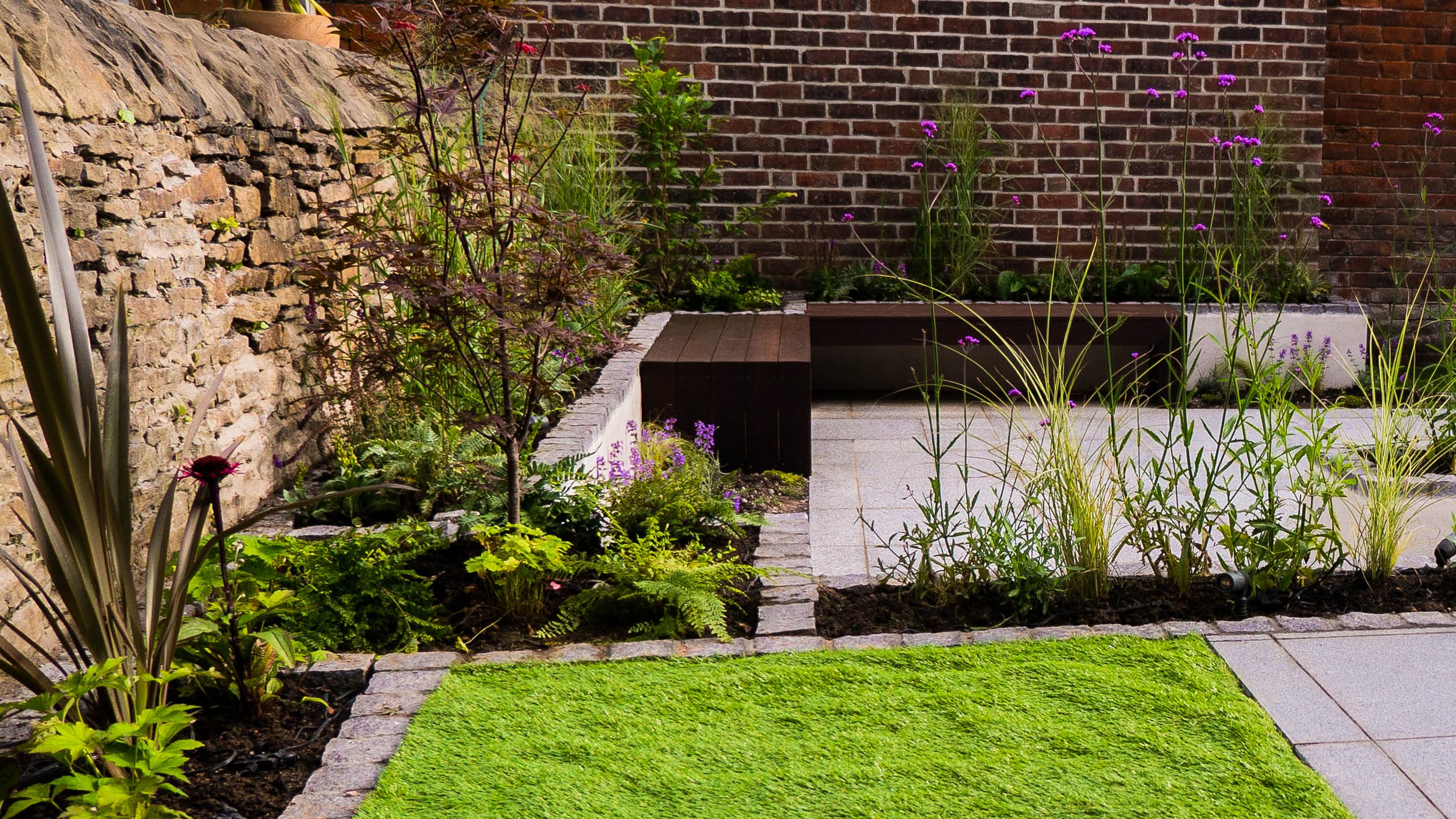 A small garden with a stone path, green grass, various flowering plants, and a brick wall backdrop.