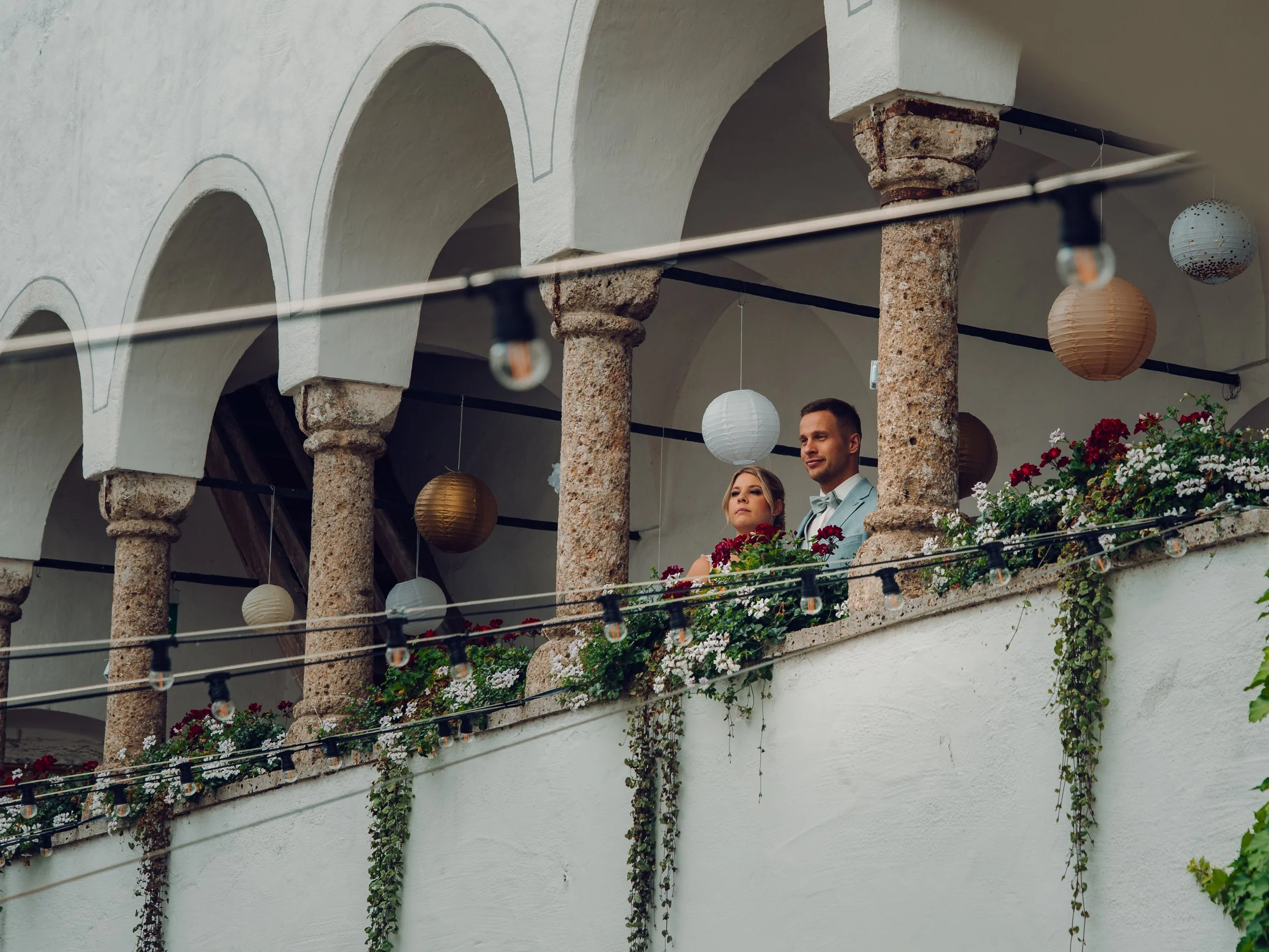 Ein Paar in festlicher Kleidung auf einer Terrassenbalustrade mit Blumen, Dachbögen im Hintergrund, dekoriert mit Papierlampions und lichterketten.