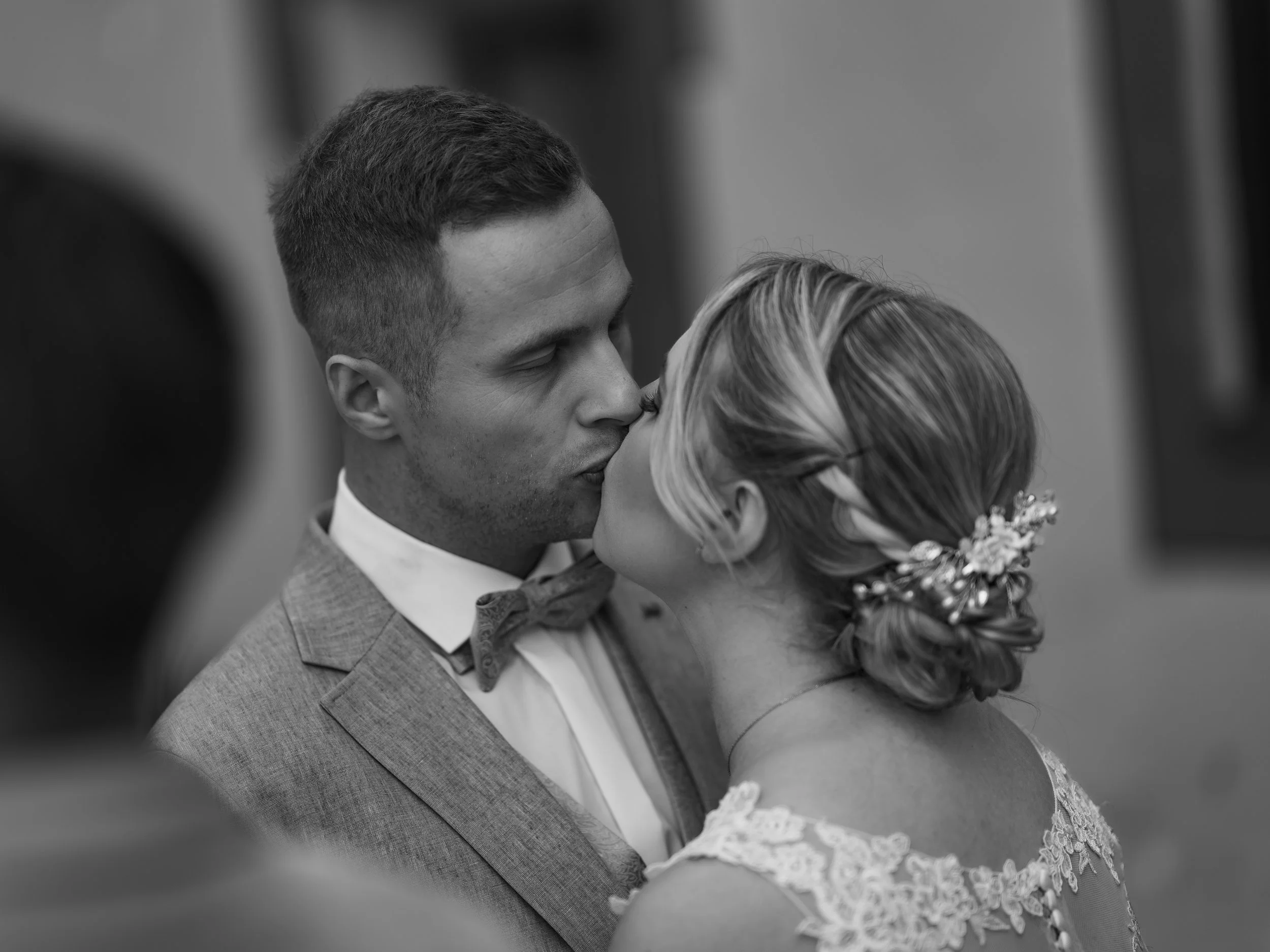 Braut und Bräutigam küssen sich bei ihrer Hochzeit, im Hintergrund sind weitere Gäste sichtbar.