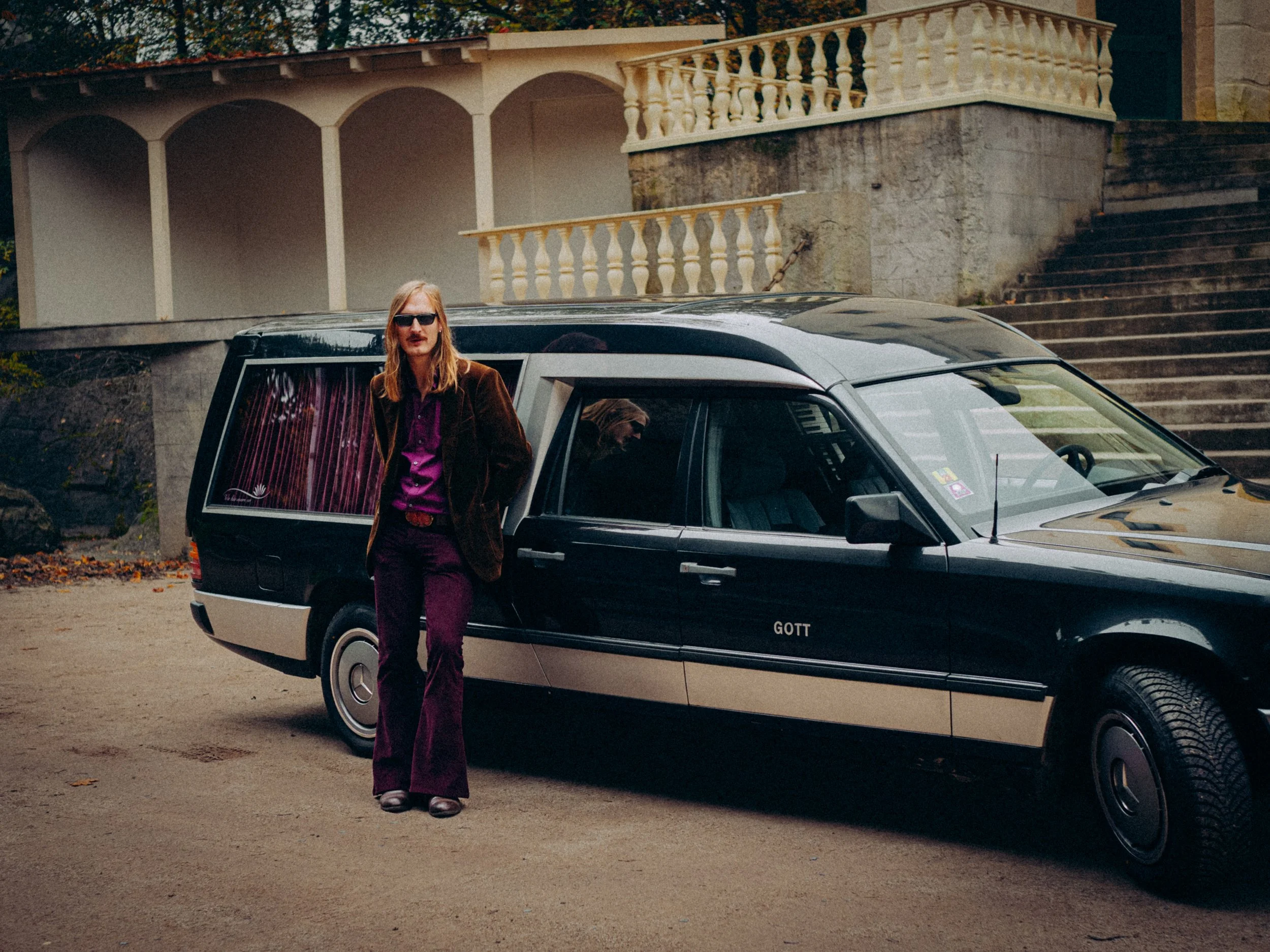 Ein Mann mit langen Haaren, Sonnenbrille, braunem Jacke und lila Outfit steht neben einem schwarzen Mercedes-Oldtimer vor einem alten Gebäude mit Treppen und Balkon.