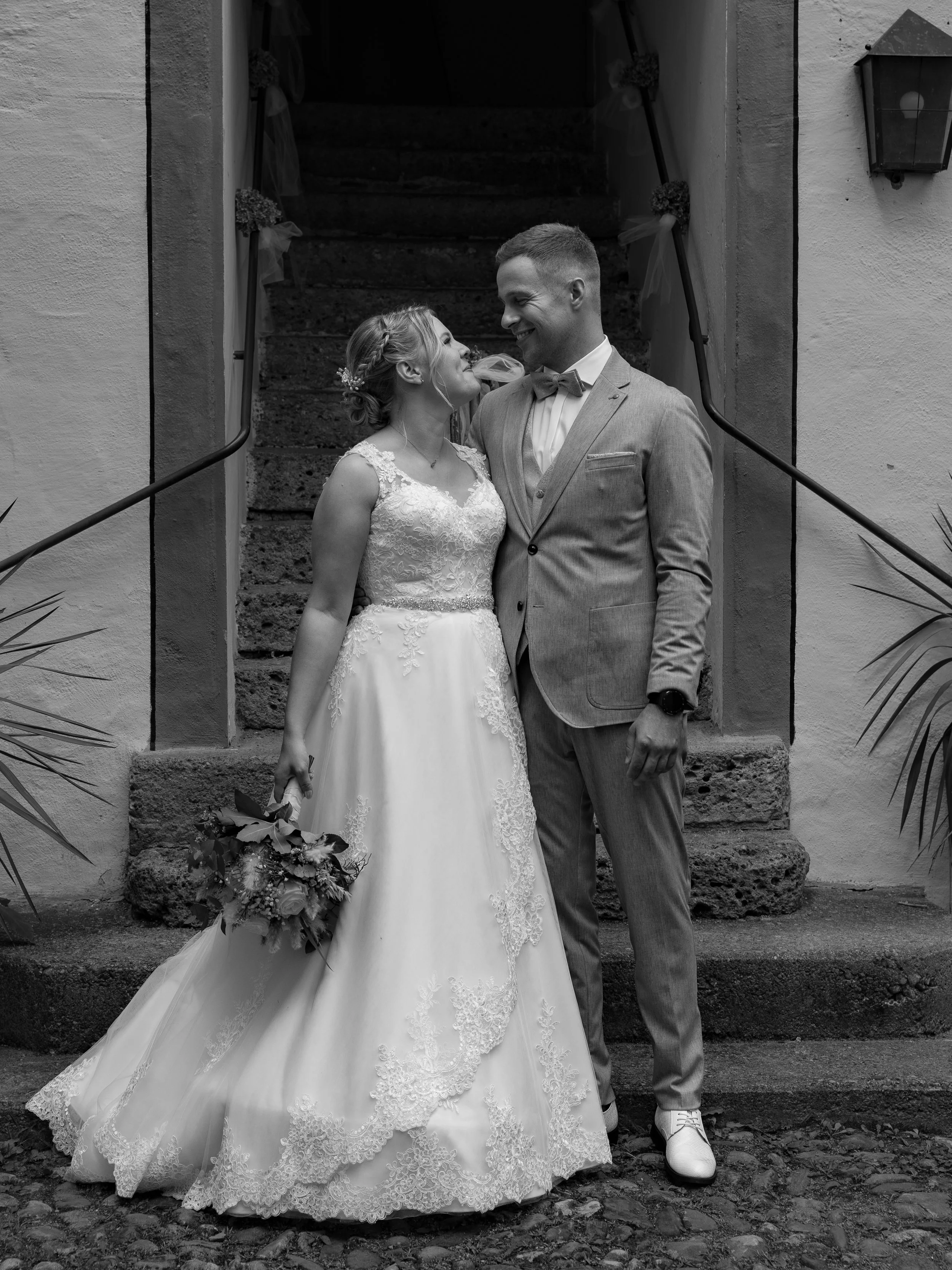 Braut und Bräutigam stehen sich an einer Treppe gegenüber, die für die Hochzeit geschmückt ist, lachend und Blick in die Augen. Die Braut trägt ein elegantes weißes Hochzeitskleid mit Blumenmuster und hält einen Blumenstrauß. Der Bräutigam trägt eine