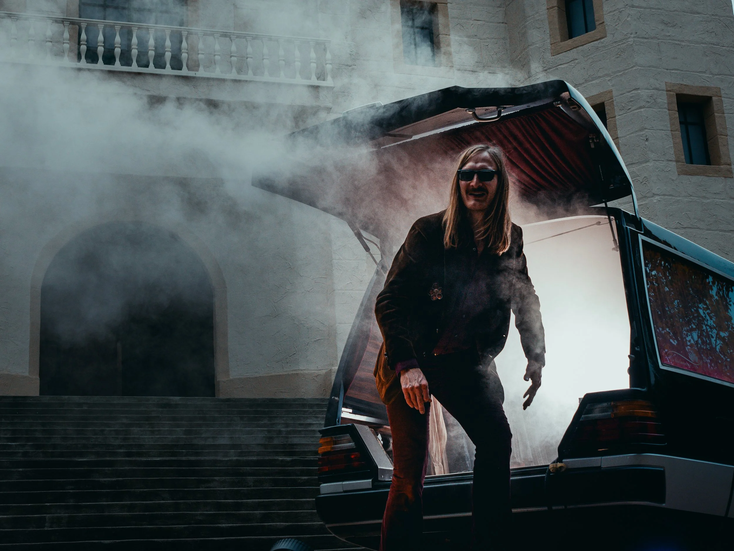 Ein Mann mit langen Haaren, Sonnenbrille und dunkler Kleidung steht in einem aufklappbaren Autokofferraum, umgeben von Rauch, vor einem historischen Gebäude mit Steintreppen und einer Balustrade.