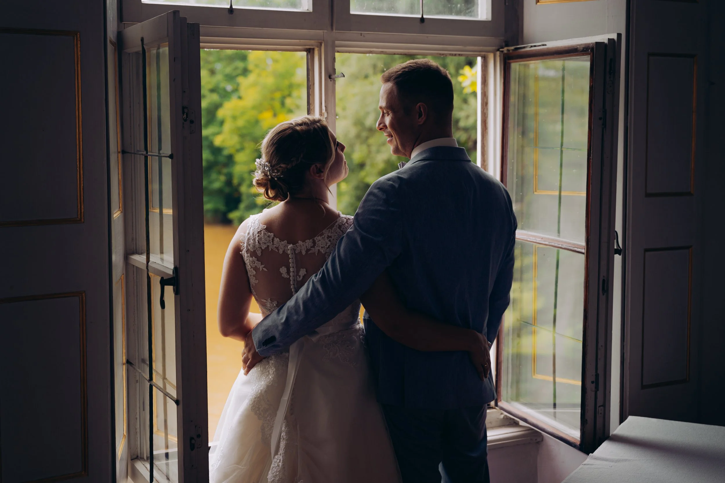 Verheiratetes Paar steht vor einem offenen Fenster, umarmt sich und blickt sich liebevoll an.