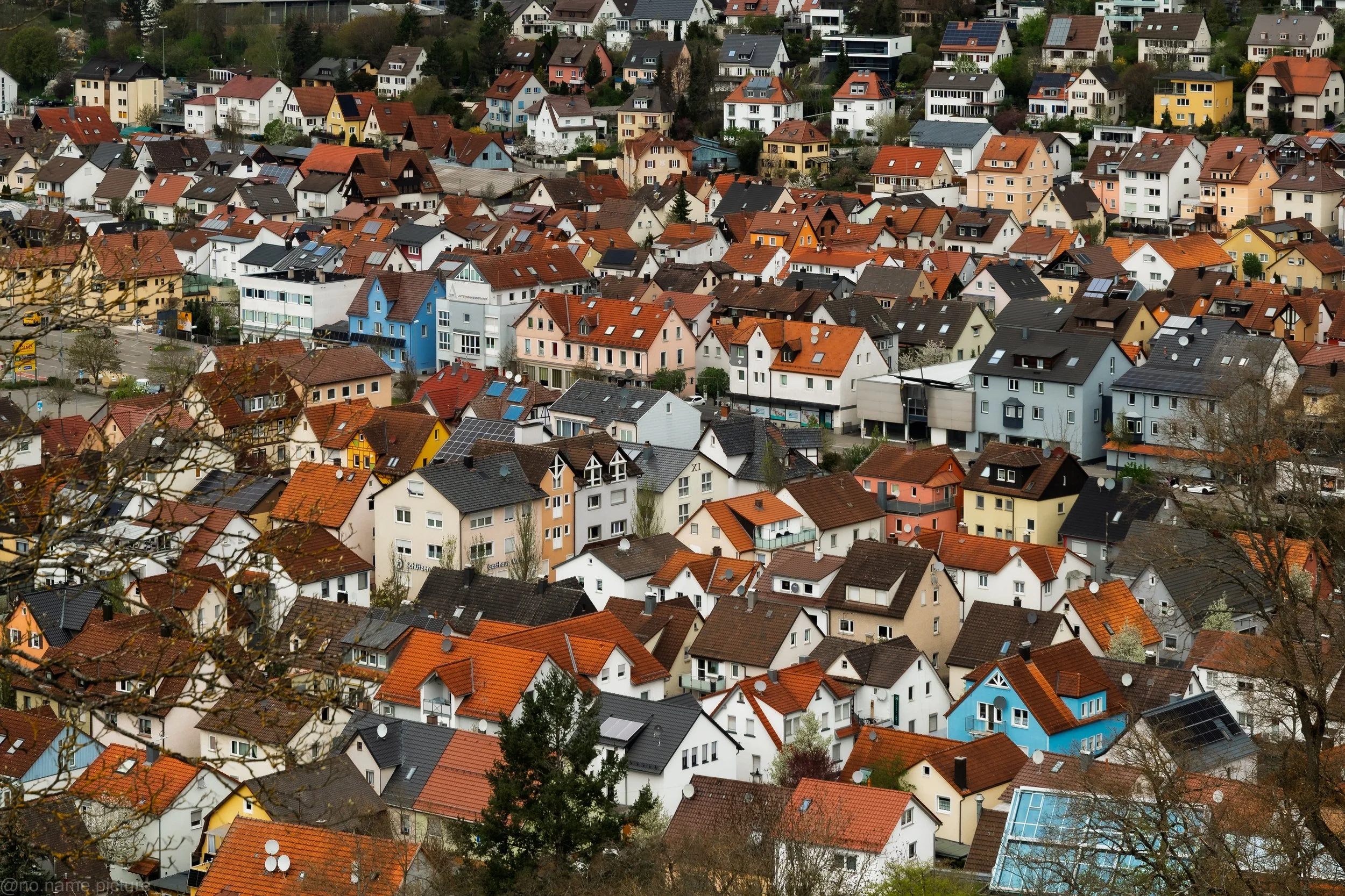 Blick auf eine hügelige Stadt mit vielen Häusern in verschiedenen Farben, darunter rot, blau, gelb und weiß, mit unterschiedlichen Dachstilen und -materialien, teilweise mit Solarpanels. Einige Bäume sind im Vordergrund erkennbar.