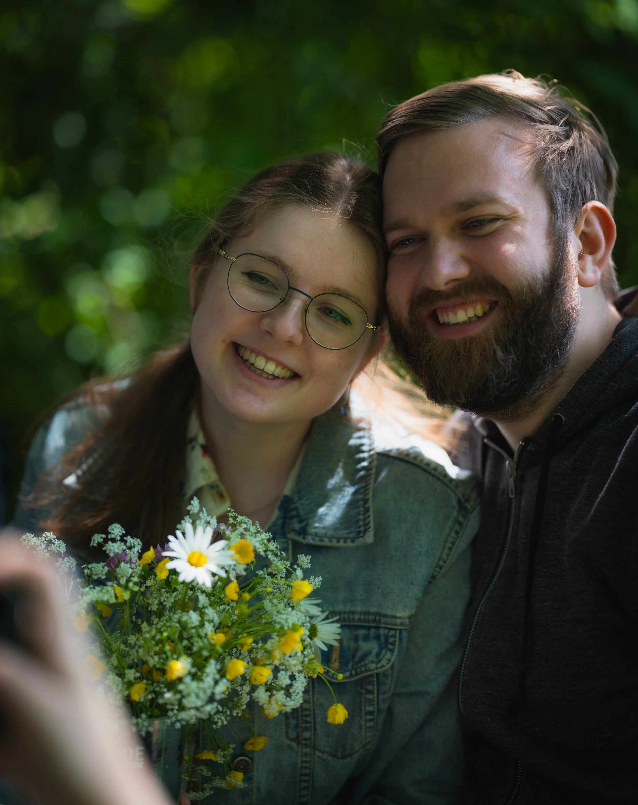Ein lachendes Paar fotografiert sich in einem Wald. Die Frau trägt eine Brille und hält einen Blumenstrauß aus Gänseblümchen und gelben Blumen.