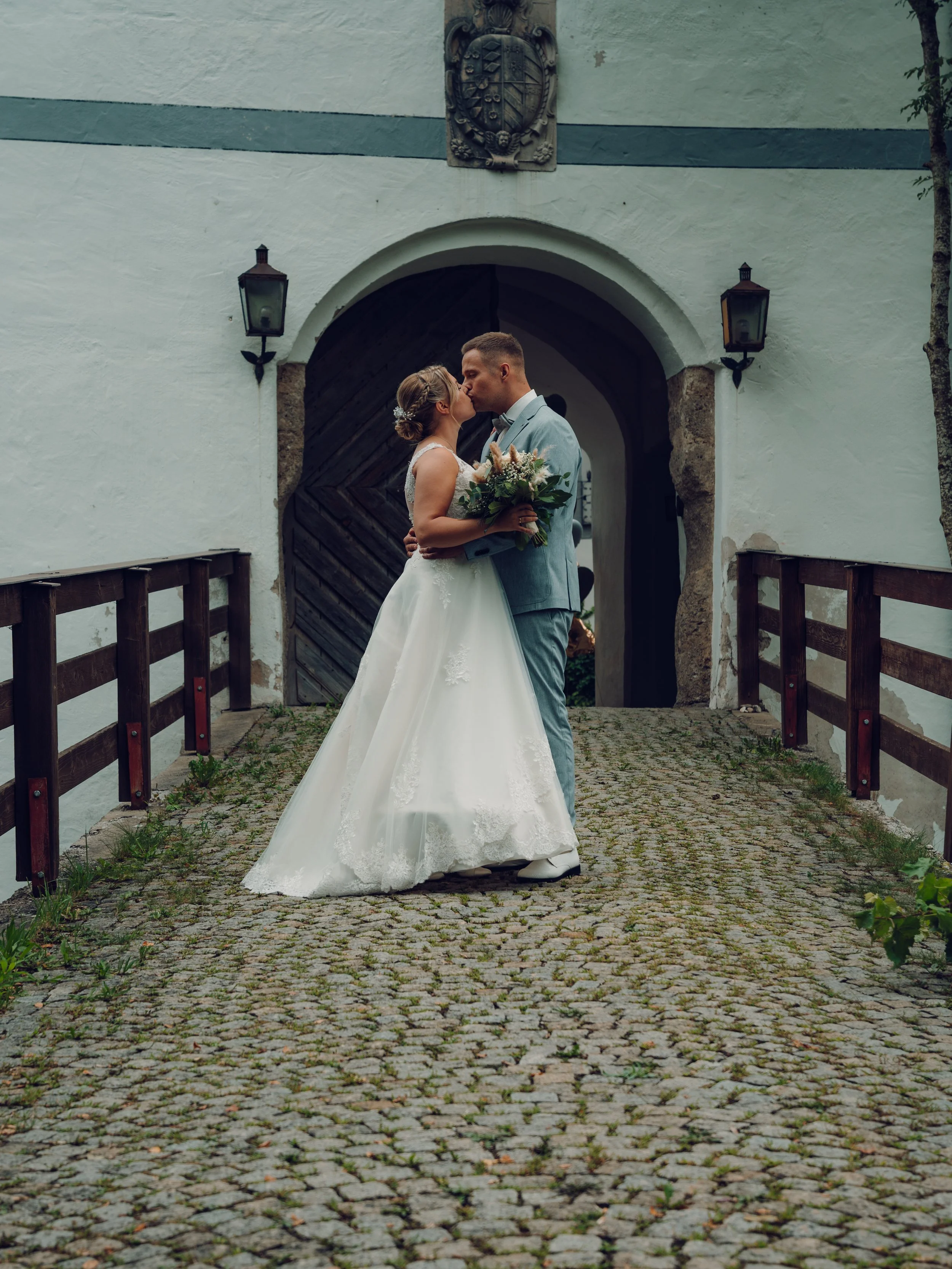 Ein Brautpaar steht sich vor einer alten, weißen Hauswand mit Holzportal und umarmt sich bei Sonnenuntergang.
