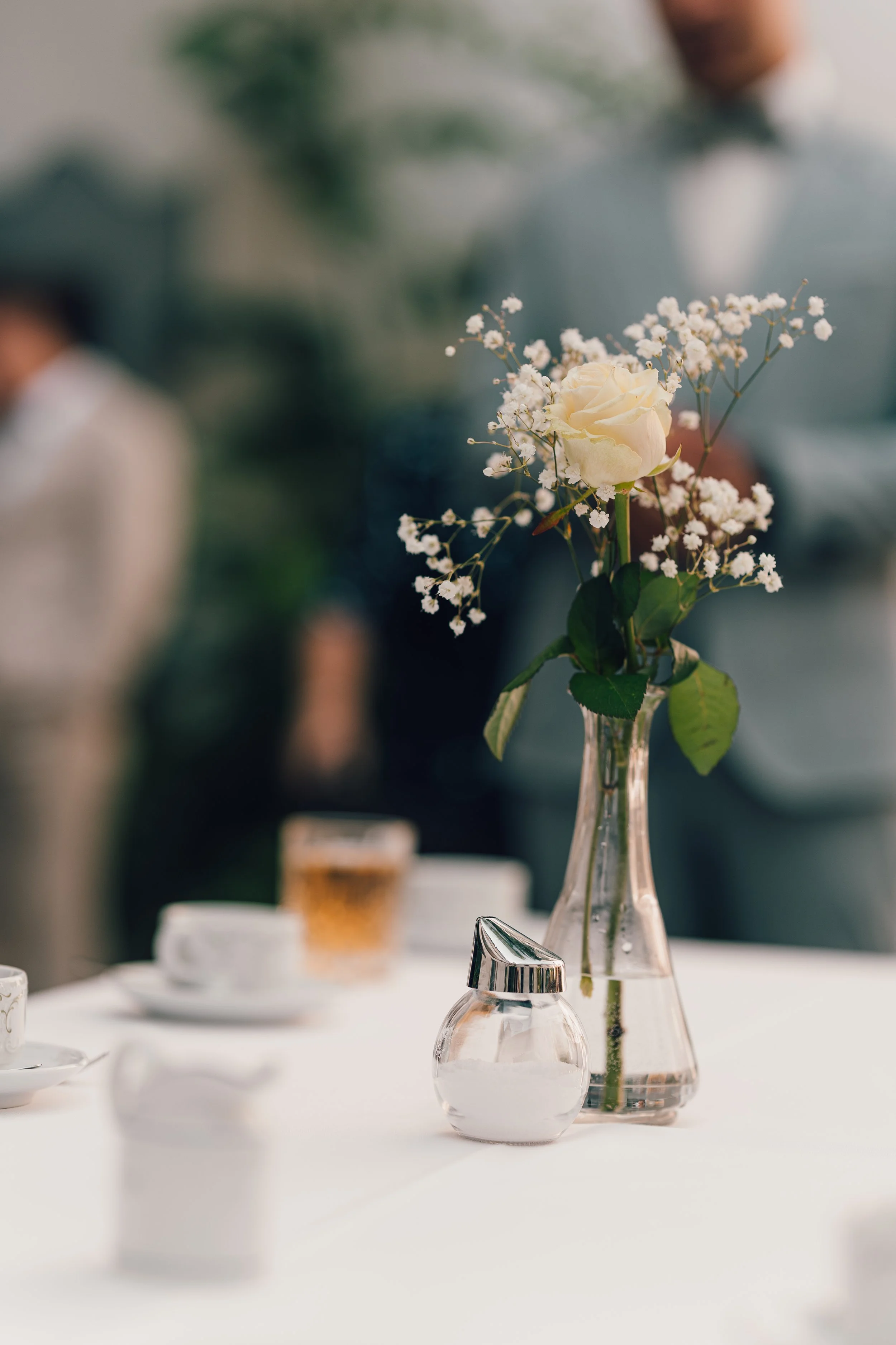 Blumenstrauß mit weißen Rosen und Gänseblümchen in einer Glasvase auf einem weißen Tisch, im Hintergrund unscharfe Menschen bei einer Veranstaltung.