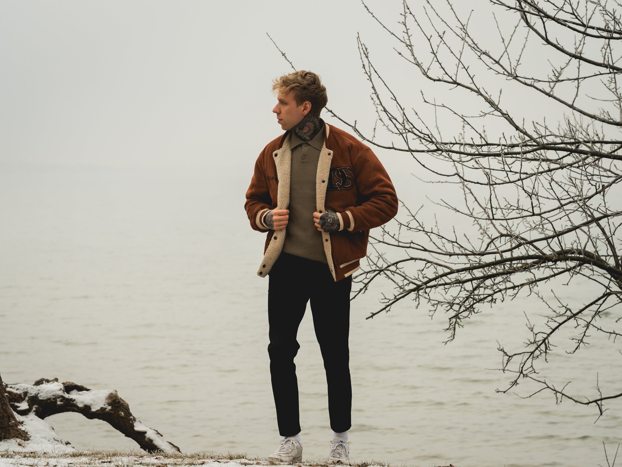 Junge steht am Ufer eines Sees bei kühlem Wetter, trägt einen braunen Jacke, grauen Pullover, schwarze Hosen und weiße Schuhe, während kahle Bäume im Hintergrund sind.
