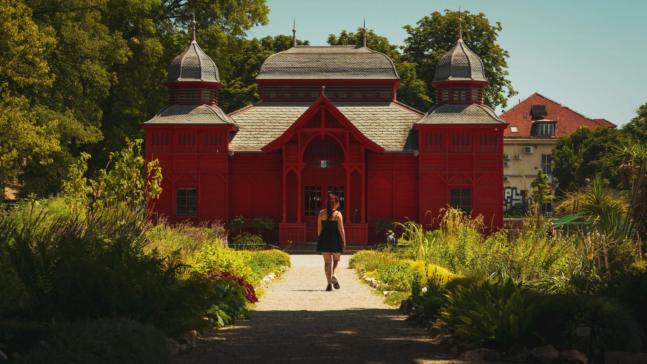 Eine junge Frau in einem schwarzen Kleid läuft einen Gartenweg entlang zu einem roten Holzhaus mit zwei Türmen, umgeben von grünen Bäumen und buntem Pflanzenbeet.