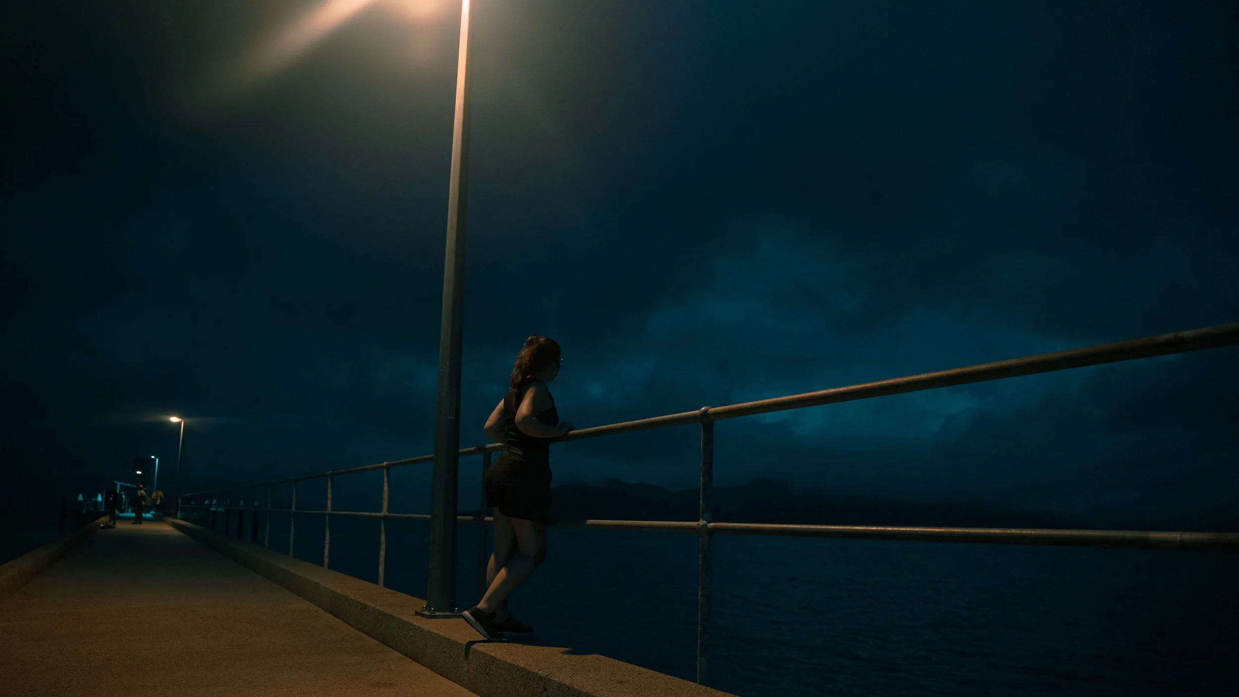 Eine Frau steht auf einem Pier bei Nacht, schaut aufs Wasser, beleuchtet von Straßenlaternen. Im Hintergrund dunkler Himmel und Meer.