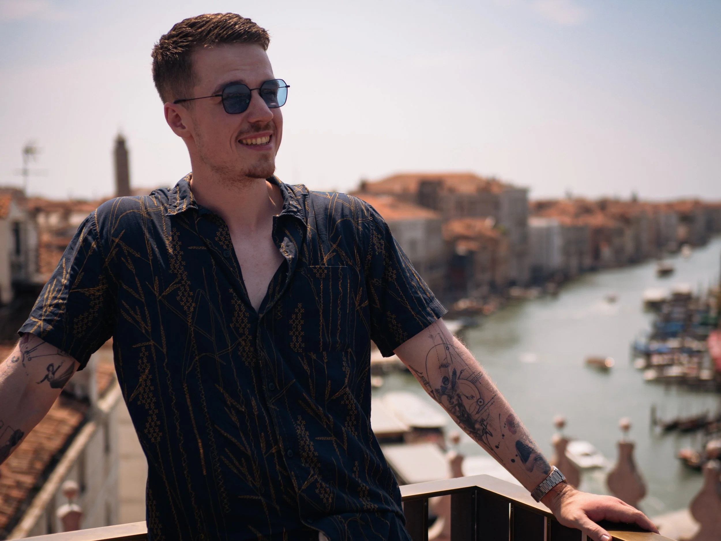 Junge Mann mit Sonnenbrille und tätowierten Armen, lächelnd, auf einer Terrasse mit Blick auf einen Kanal und historische Gebäude in Venedig.