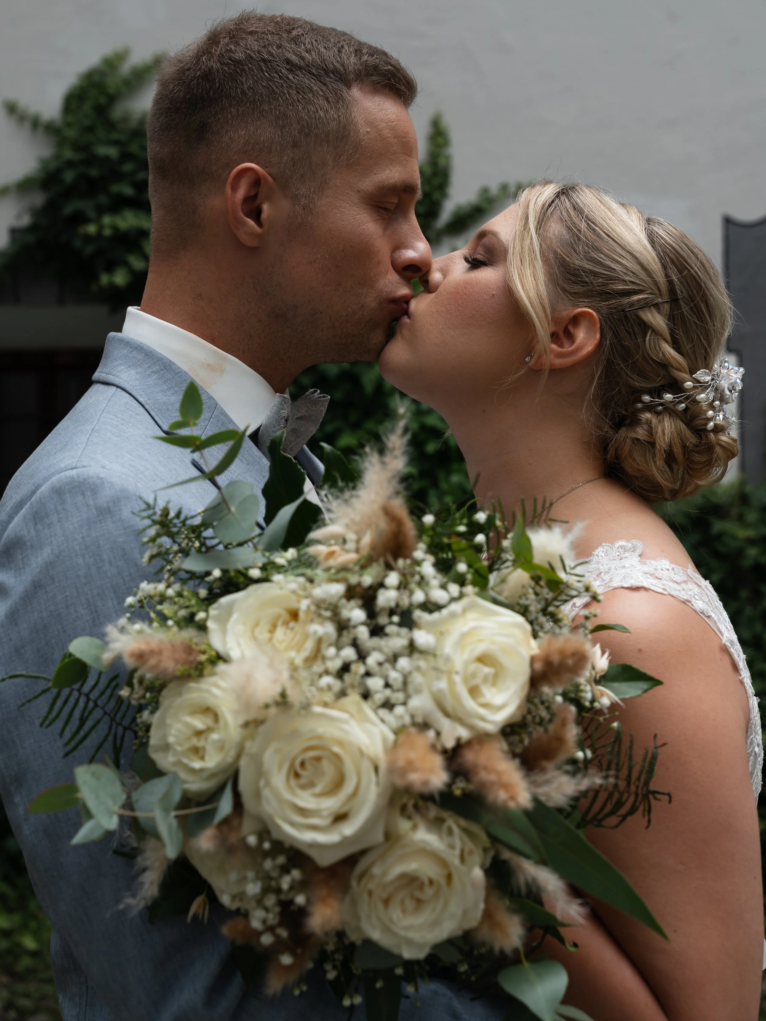 Ein Hochzeitsbild mit einem Brautpaar, das sich küsst, während die Braut einen weißen Blumenschmuck hält.