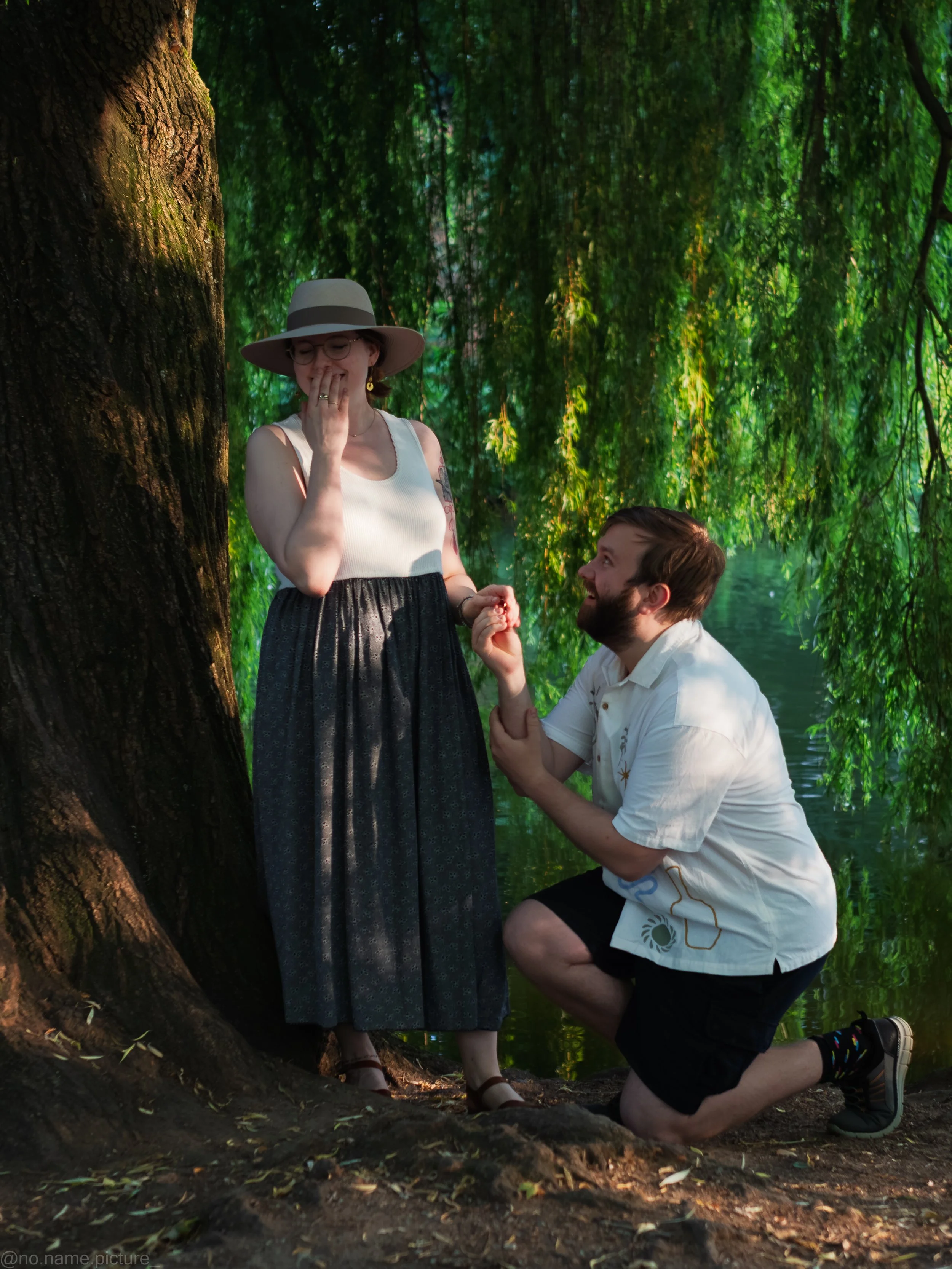 Ein Mann macht seiner Frau auf einem Stein am Wasser einen Heiratsantrag. Sie lachen und freuen sich.