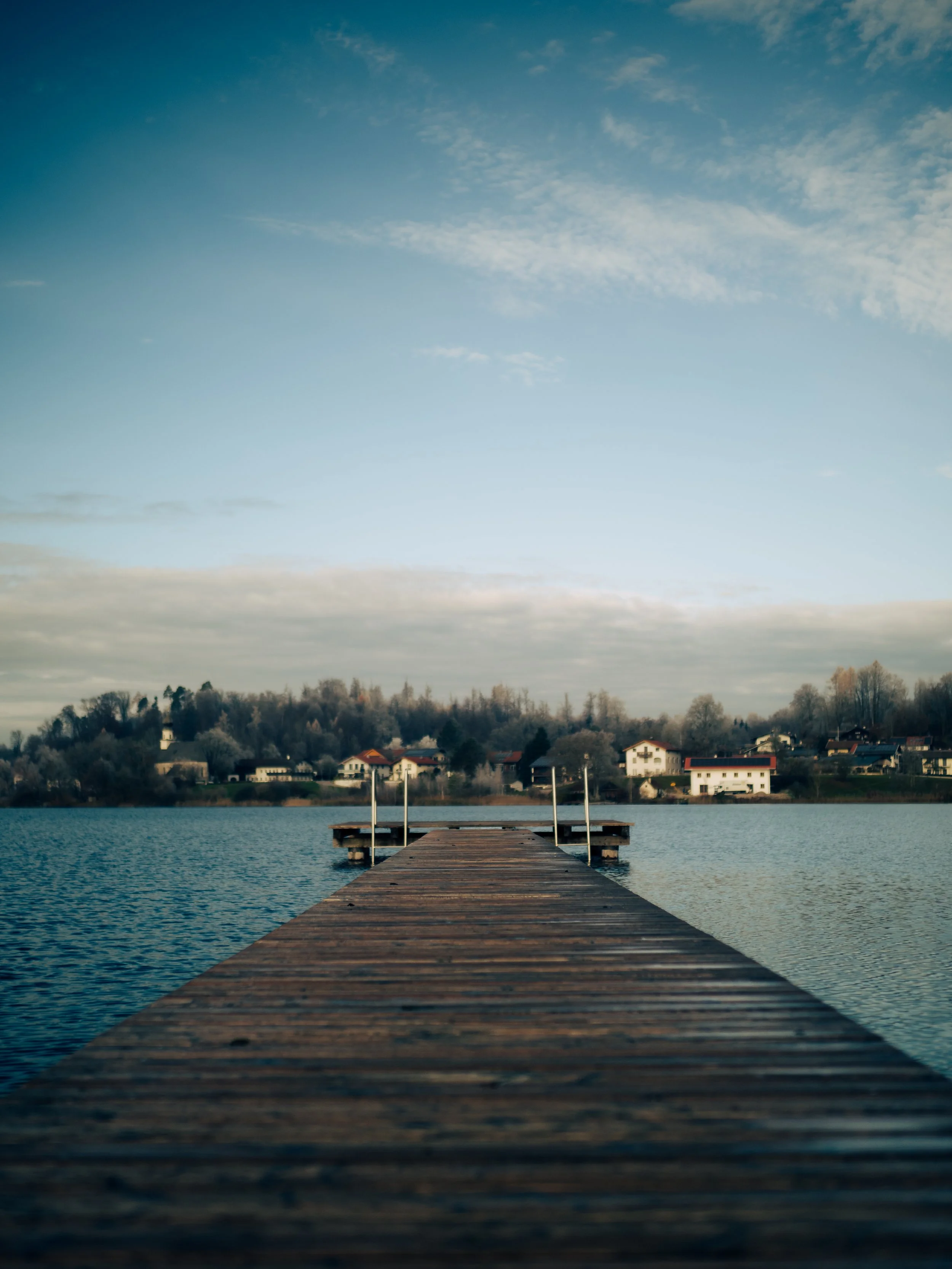 Ein Holzsteg führt zu einem Wasser, mit Häusern und Bäumen im Hintergrund. Der Himmel ist teilweise bewölkt.
