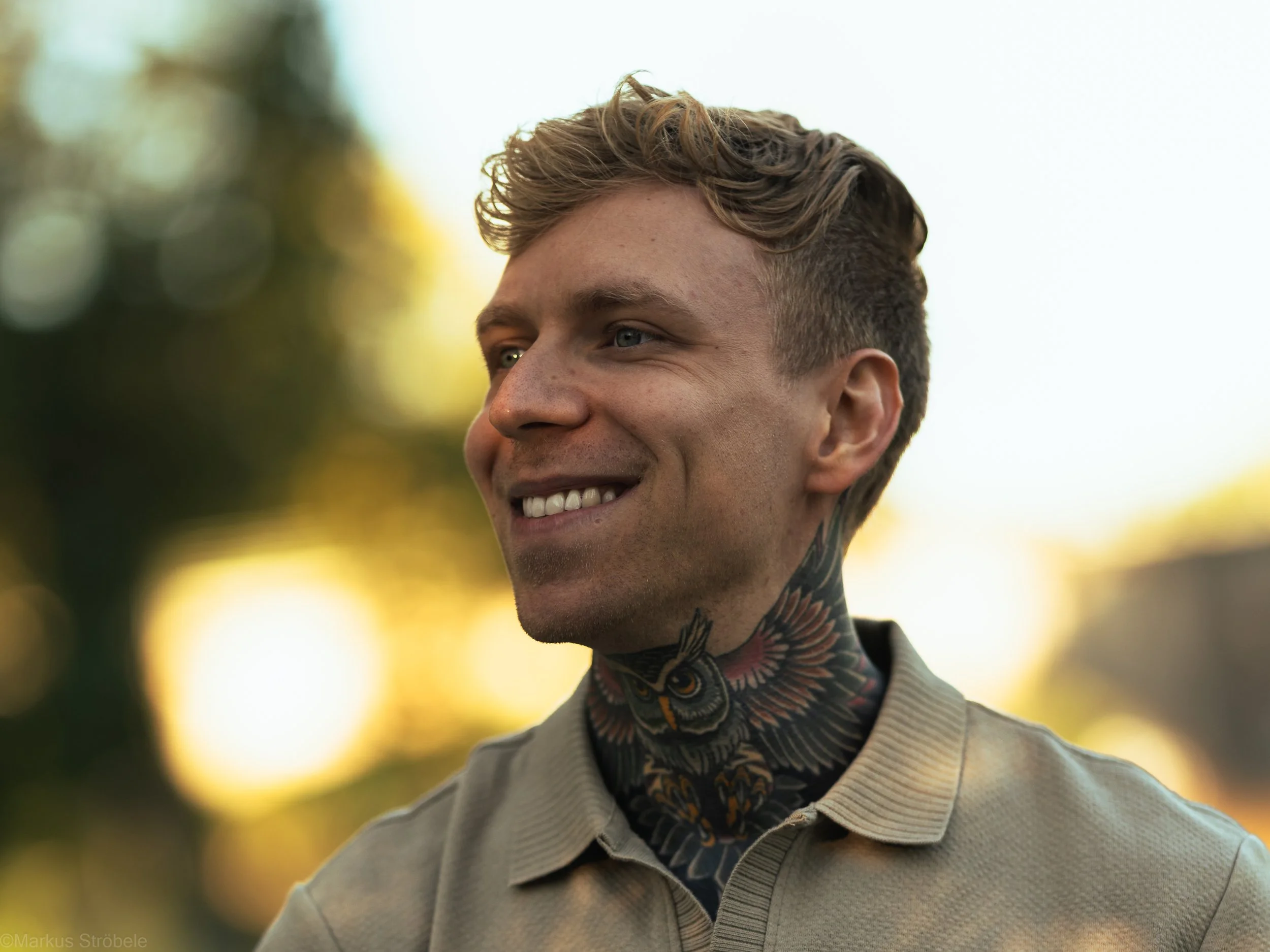 Lächelnder junger Mann mit kurzen, lockigen Haaren und Tätowierungen am Hals, im Freien bei Sonnenuntergang.
