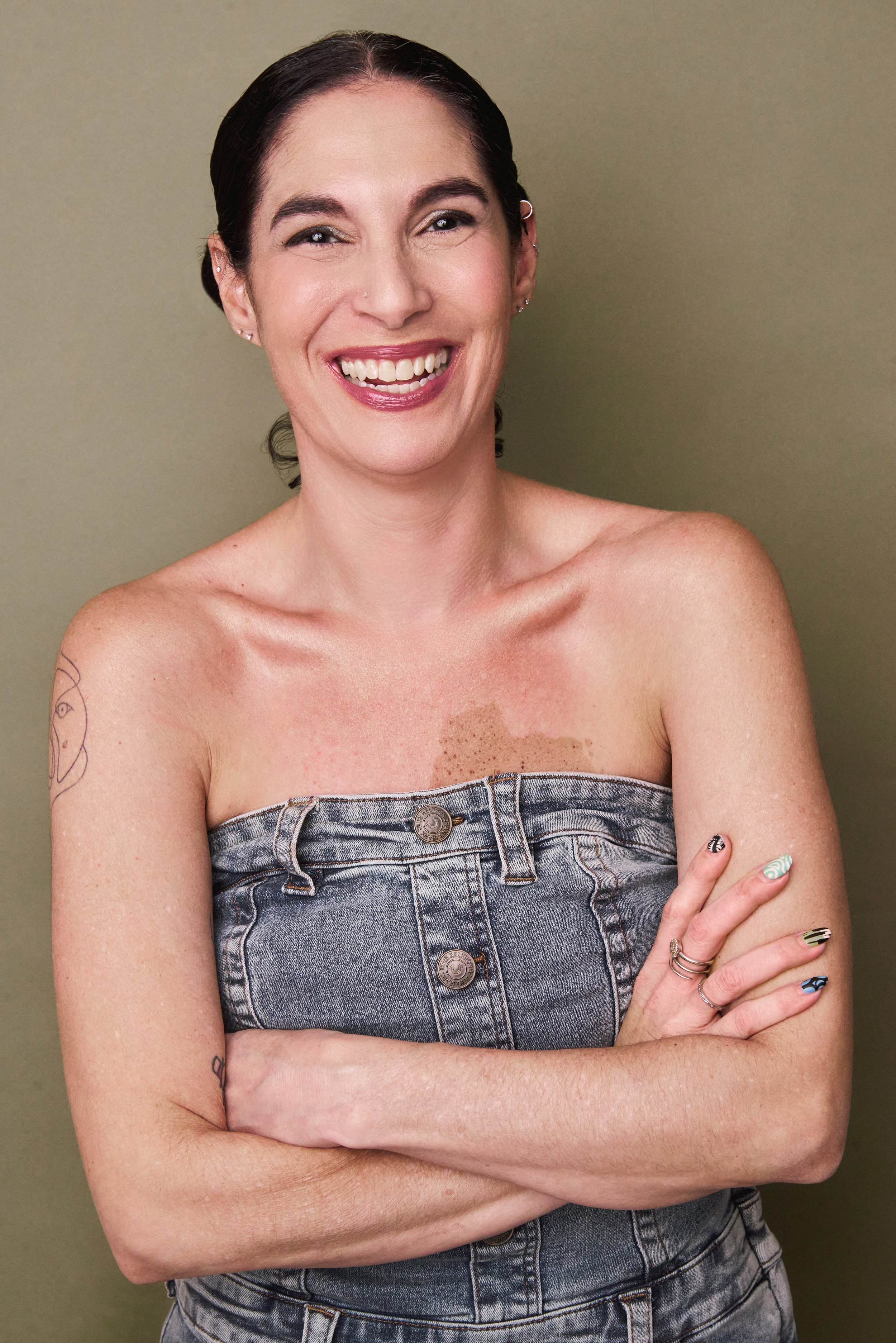 A woman with dark hair in a low bun, smiling with arms crossed, wearing a strapless denim top, showcasing tattoos on her arms and chest, against a plain background.