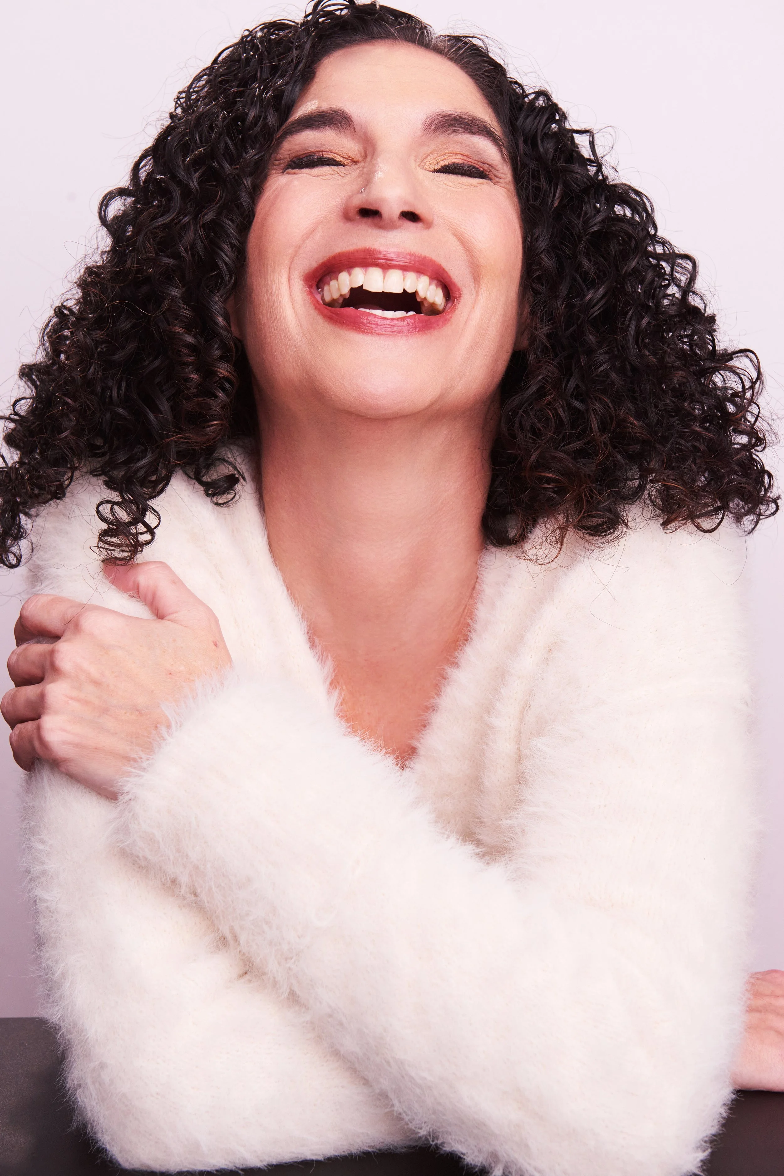 A woman with curly dark hair laughing and smiling with her eyes closed, wearing a fluffy white sweater.