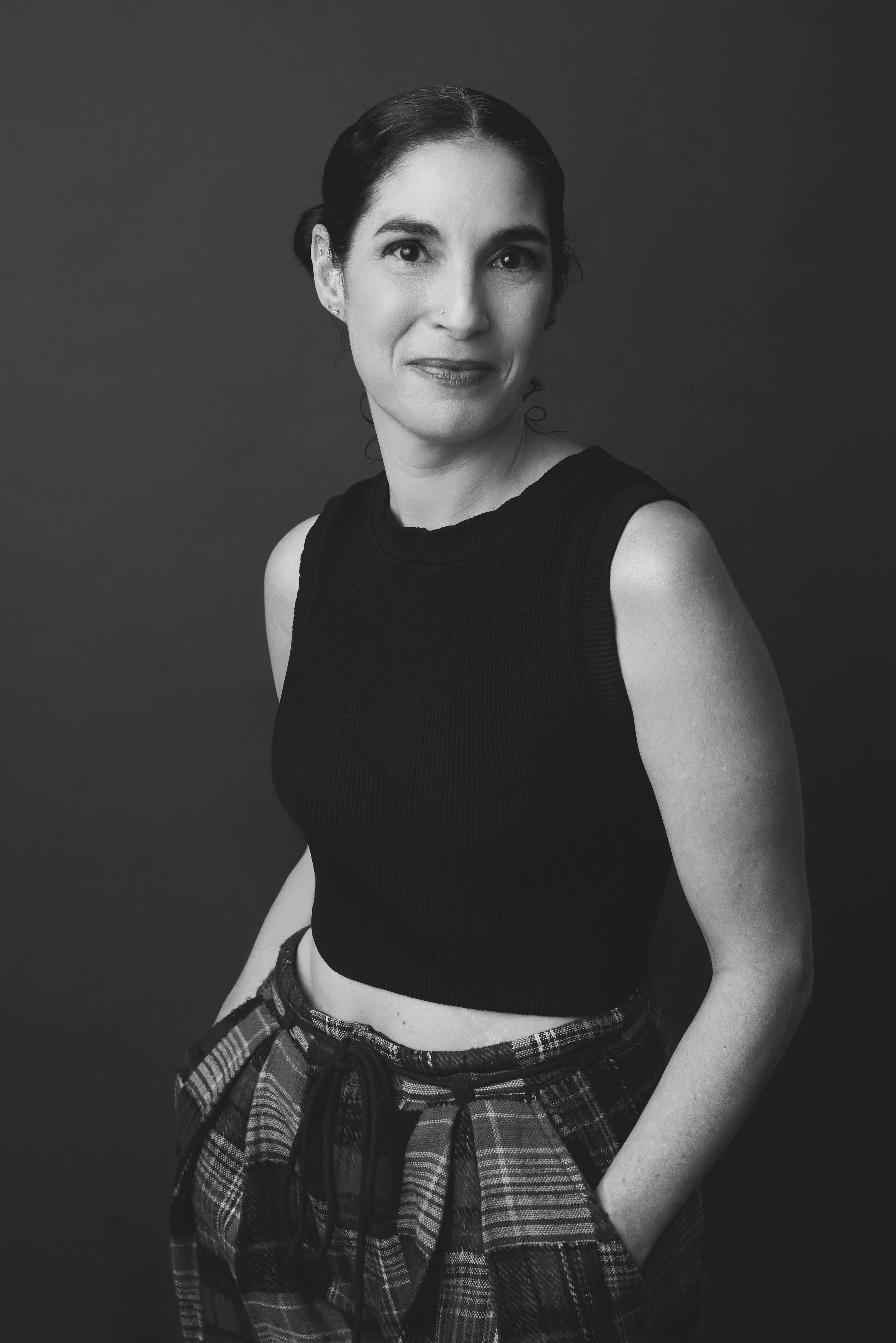 Black and white portrait of a woman with dark hair pulled back, wearing a sleeveless black top and plaid pants, standing against a plain dark background.