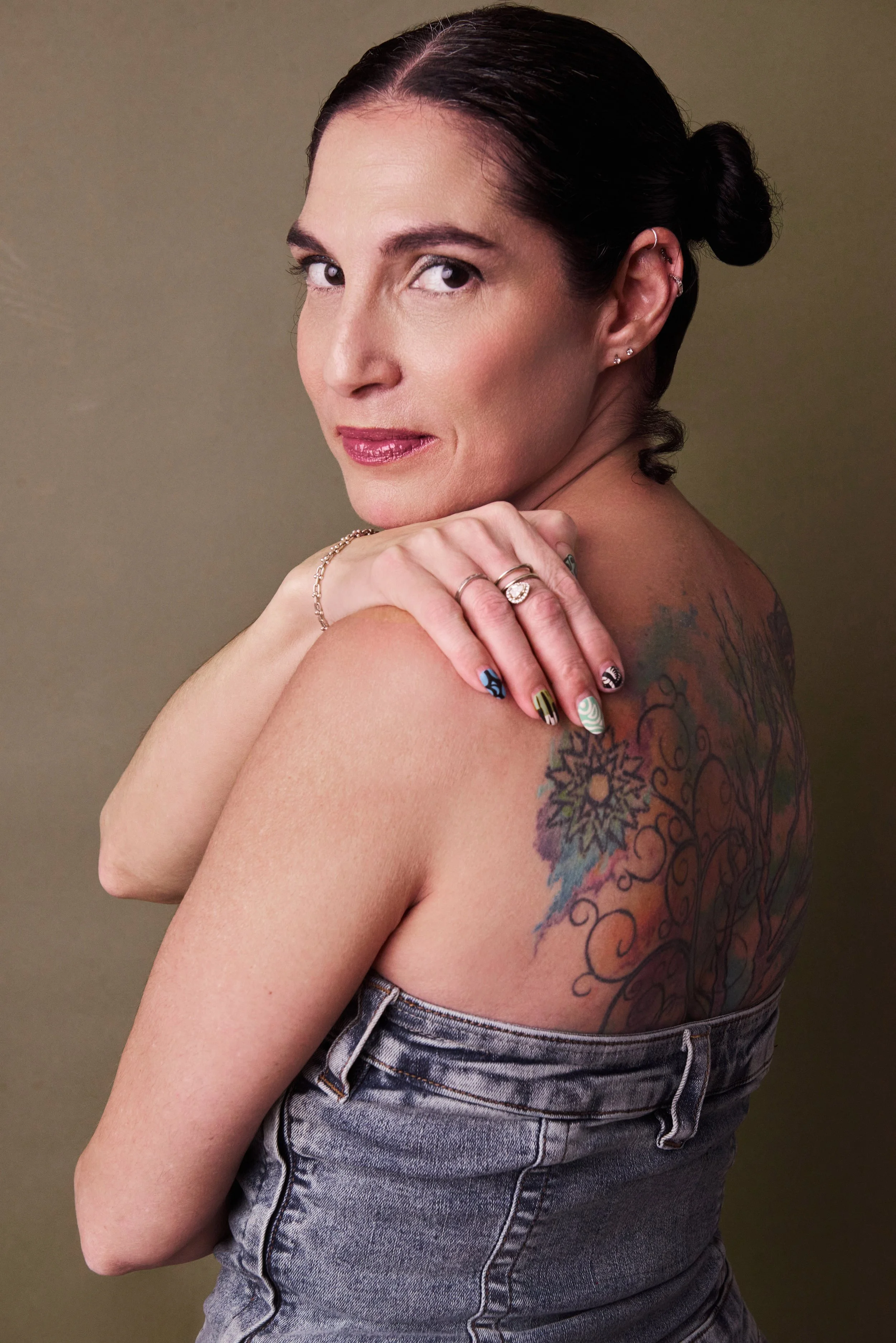 A woman with dark hair in a bun showing her back tattoo, wearing gray denim jeans, jewelry, and colorful nail polish, against a plain background.
