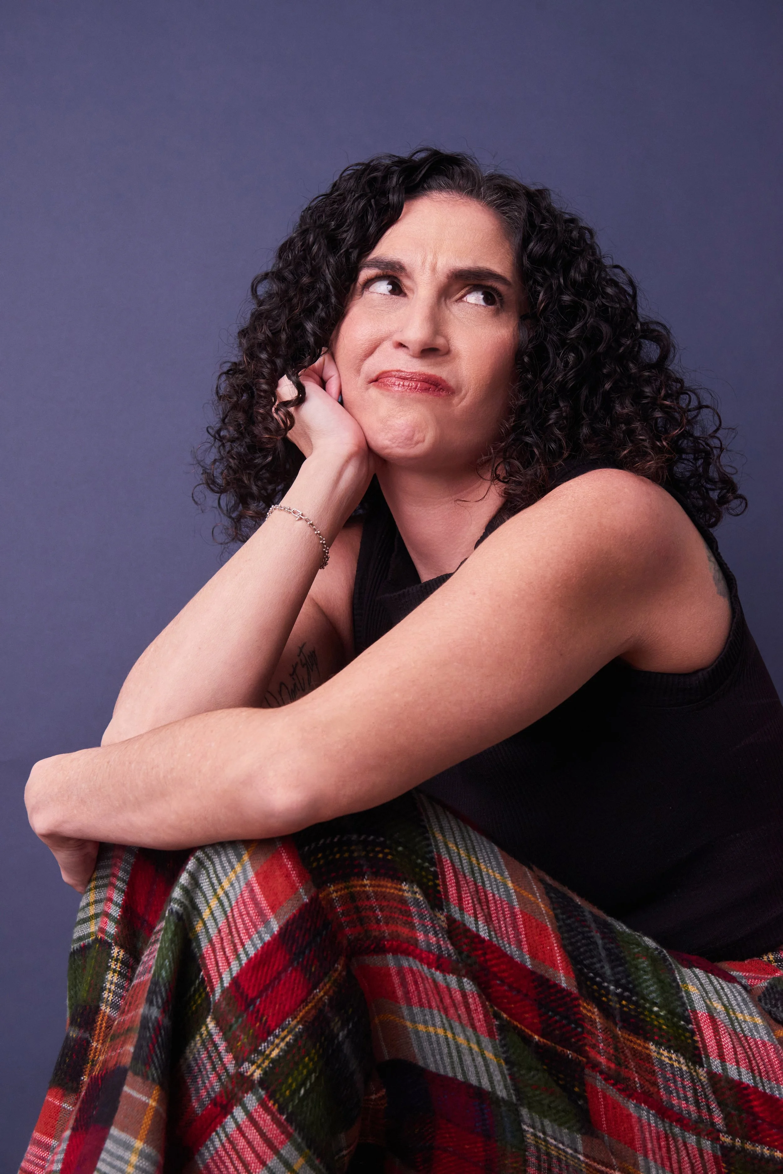 Woman with curly dark hair, resting her chin on her hand, looking puzzled, wearing a black sleeveless top and plaid pants against a gray background.