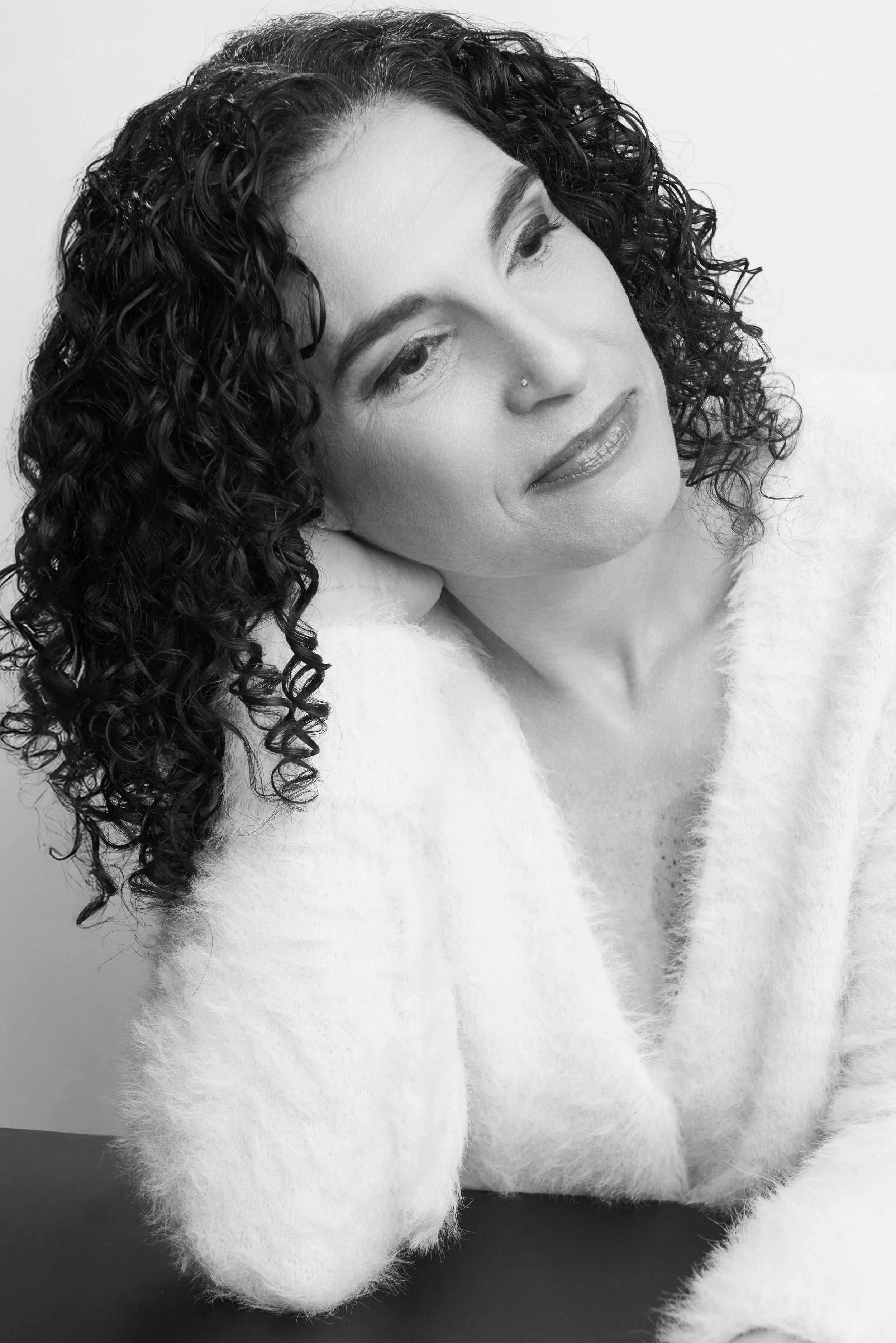 A woman with curly hair, wearing a soft fuzzy sweater, rests her head on her hand in a thoughtful pose in black and white.
