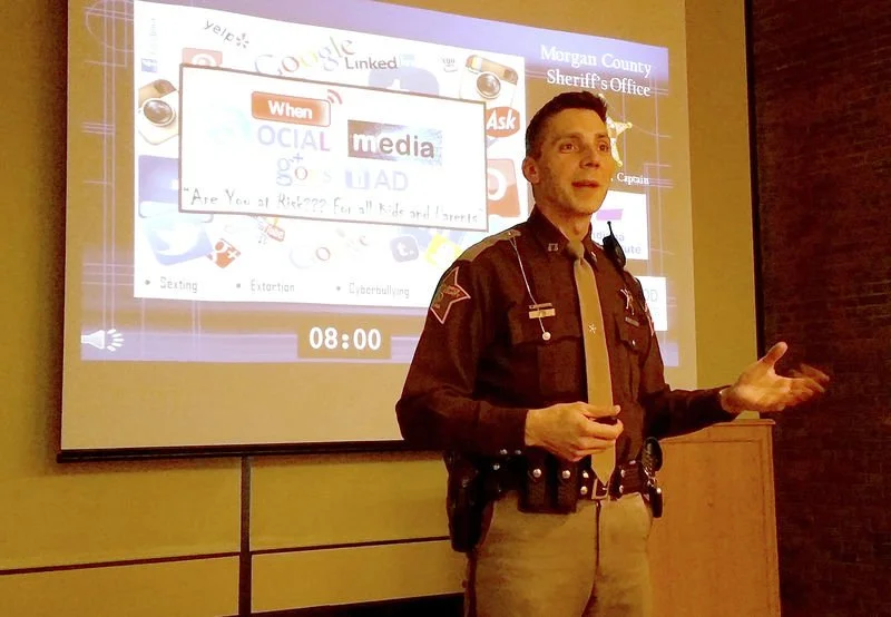 A uniformed officer giving a presentation in front of a large projector screen about social media awareness. The screen displays various social media icons, a clock showing 08:00, and text related to social media risks for kids and teens.