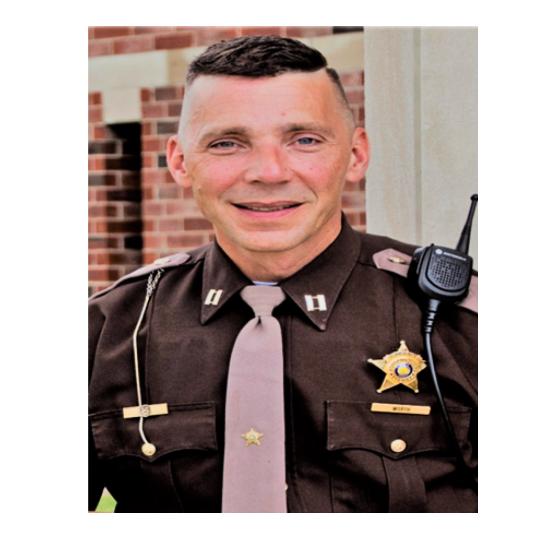 A smiling male police officer in uniform with a badge, a tie, and a microphone on his shoulder, standing outside near a brick building.