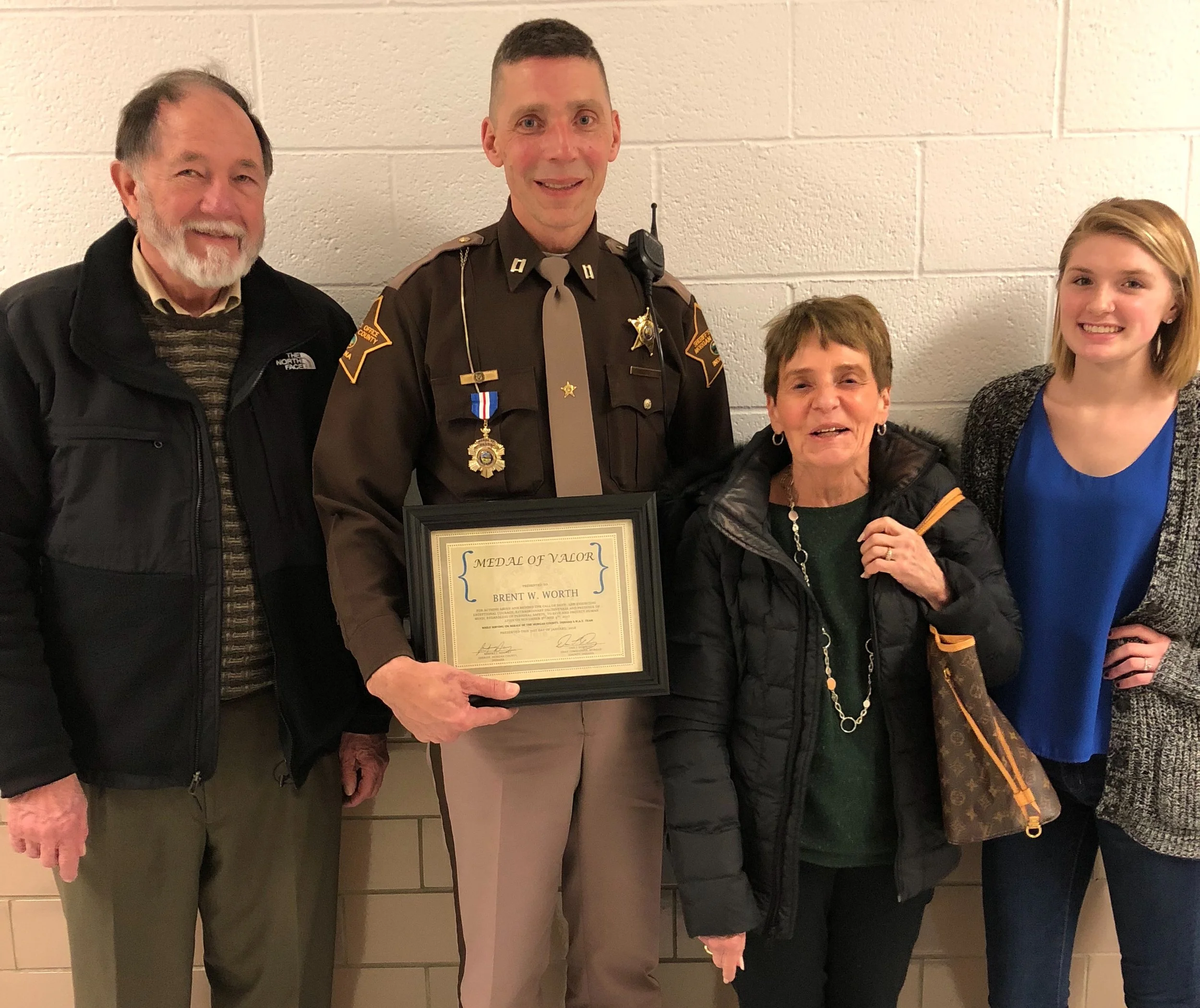 Group of five people standing against a wall, with a police officer in the center holding a framed medal of valor award.