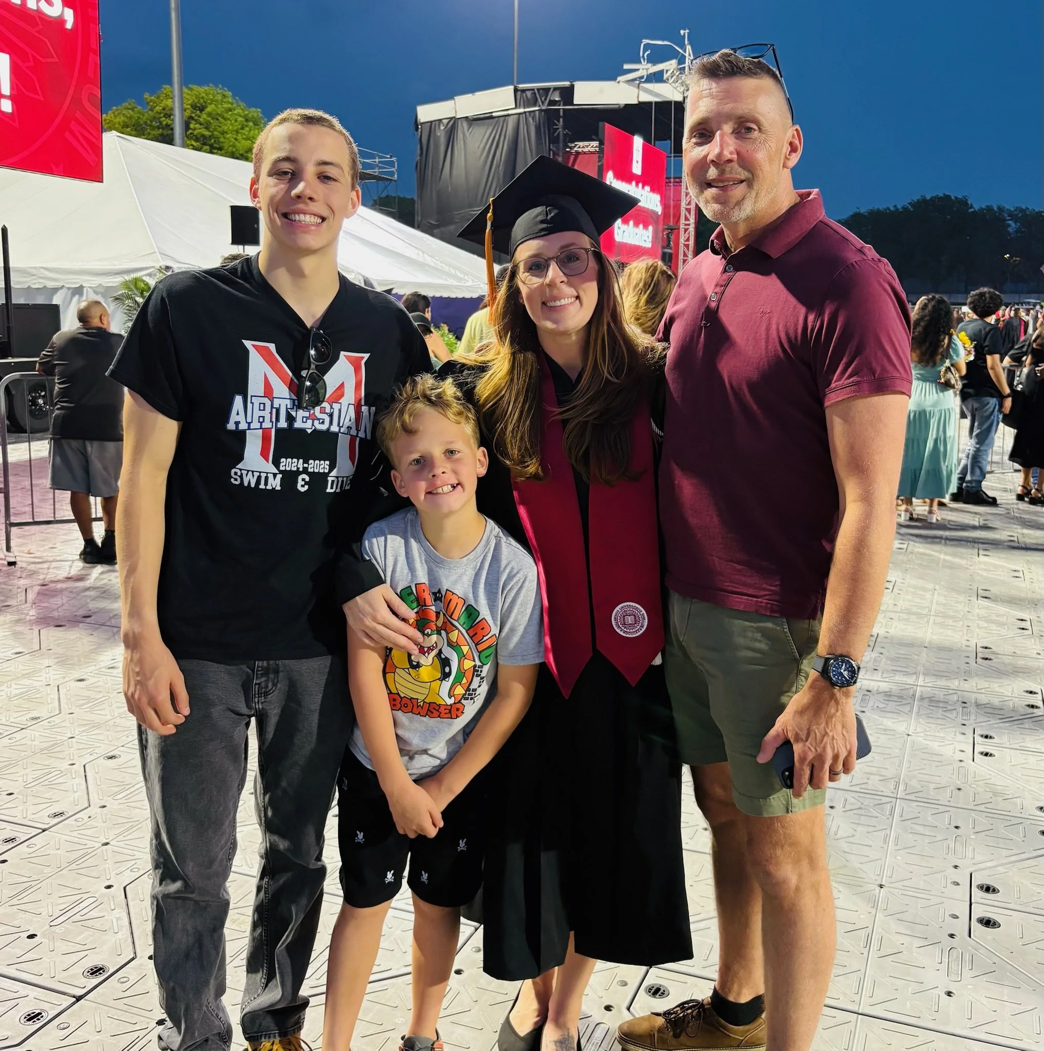 A family celebrating a graduation outdoors at night, with a woman in a cap and gown surrounded by three males, all smiling, with tents and a stage in the background.