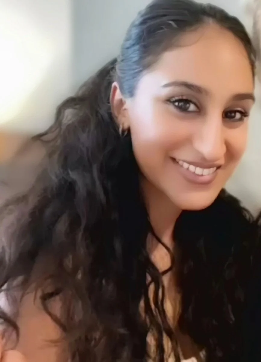 Smiling woman with long, wavy dark hair wearing earrings and a necklace, indoors.