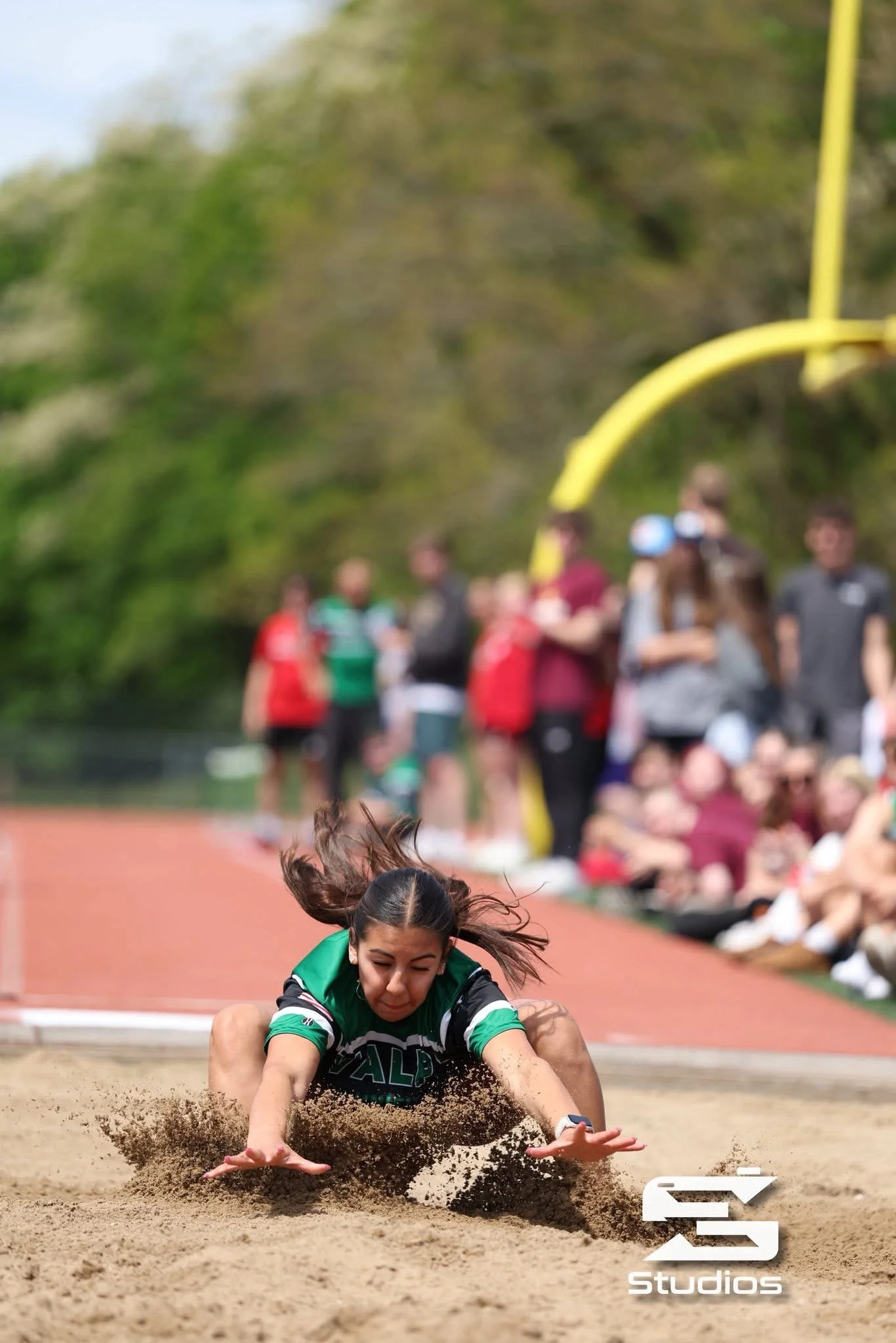 A female athlete with dark hair in a ponytail diving headfirst into a sandpit during a track and field event, with a group of people watching in the background.