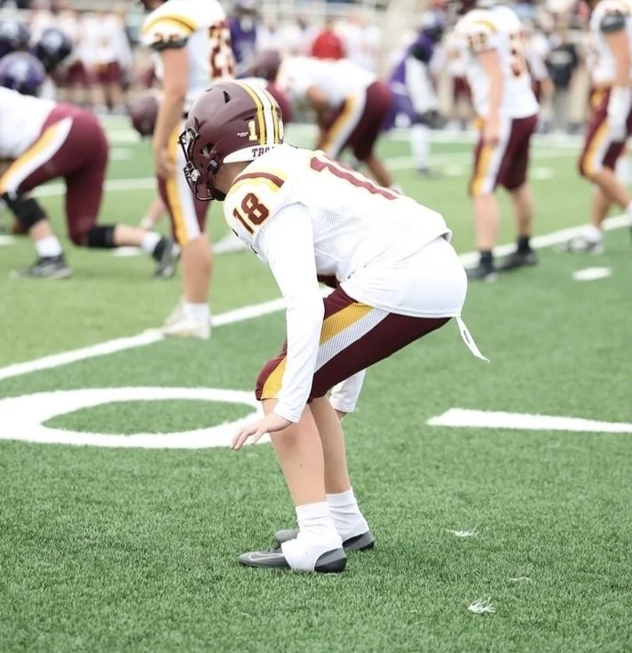 A young football player on a green field, bent down and ready to play, wearing a helmet and jersey with the number 16, surrounded by other players in similar uniforms.