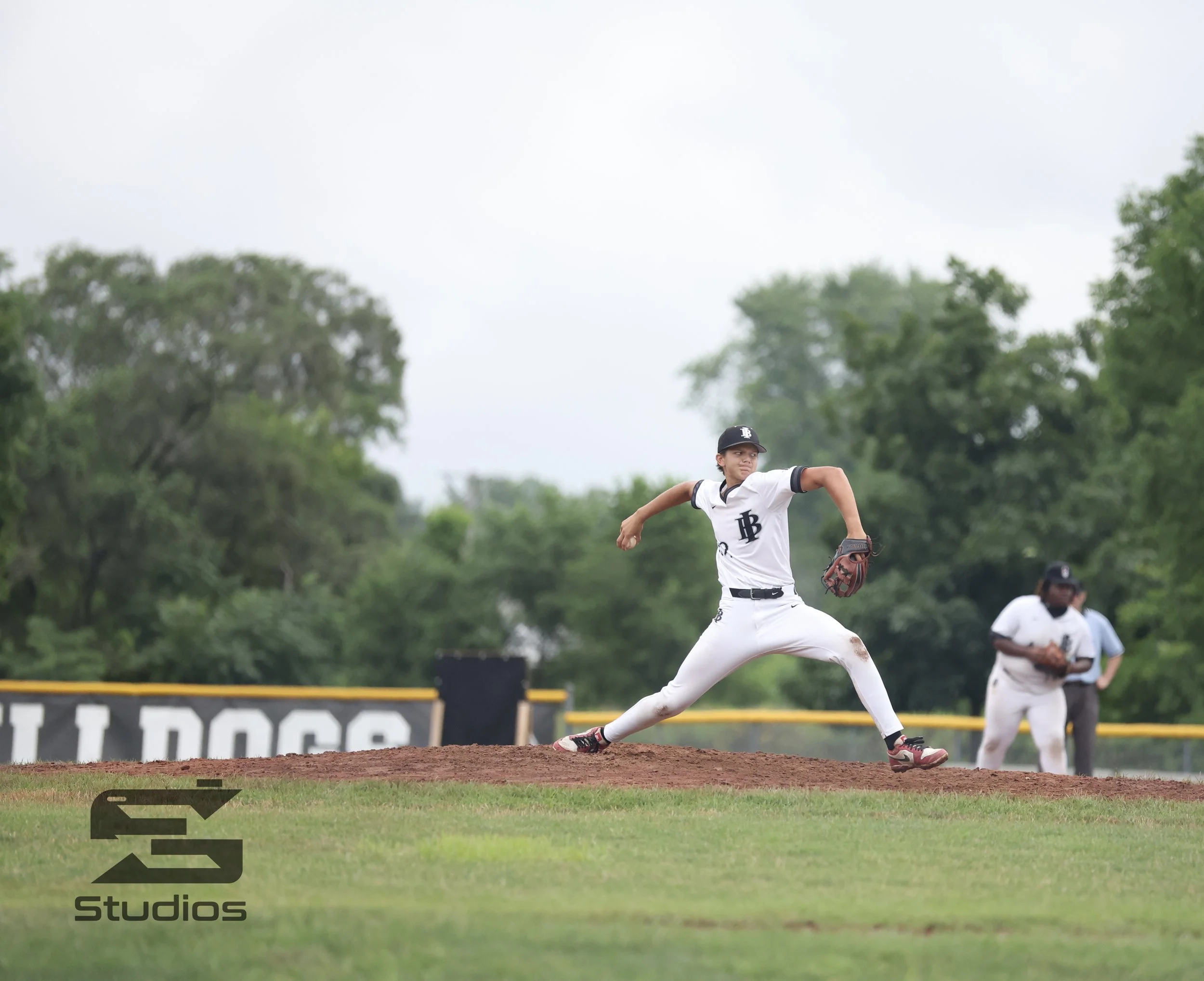 A baseball pitcher in a white uniform with black accents on the mound preparing to pitch, with a second player and an umpire in the background on a baseball field surrounded by green trees.