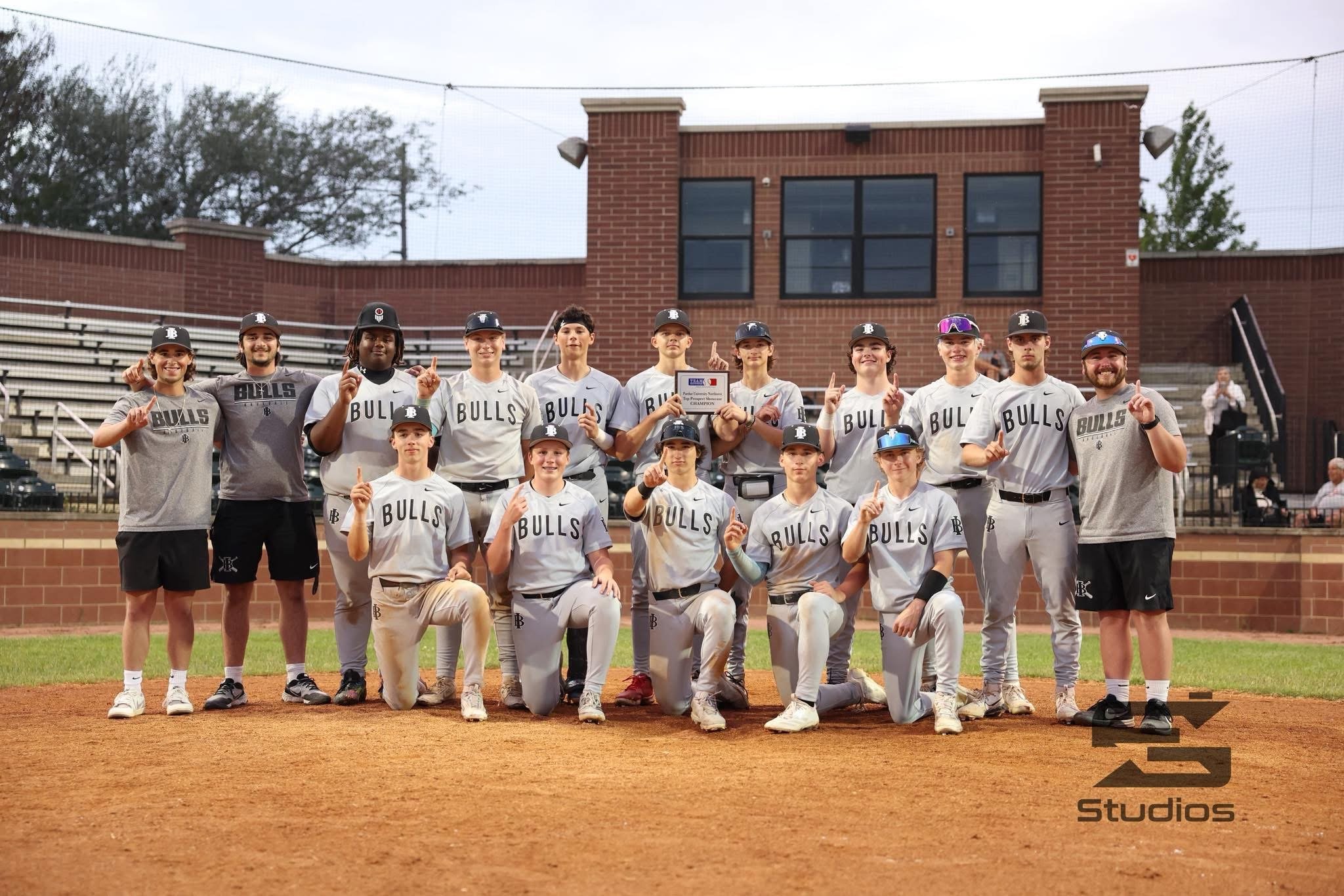 A baseball team of 14 players and coaches posing on a baseball field, holding a championship plaque and making victory signs with their fingers. They are wearing gray and black uniforms with 'BULLS' written on the front.