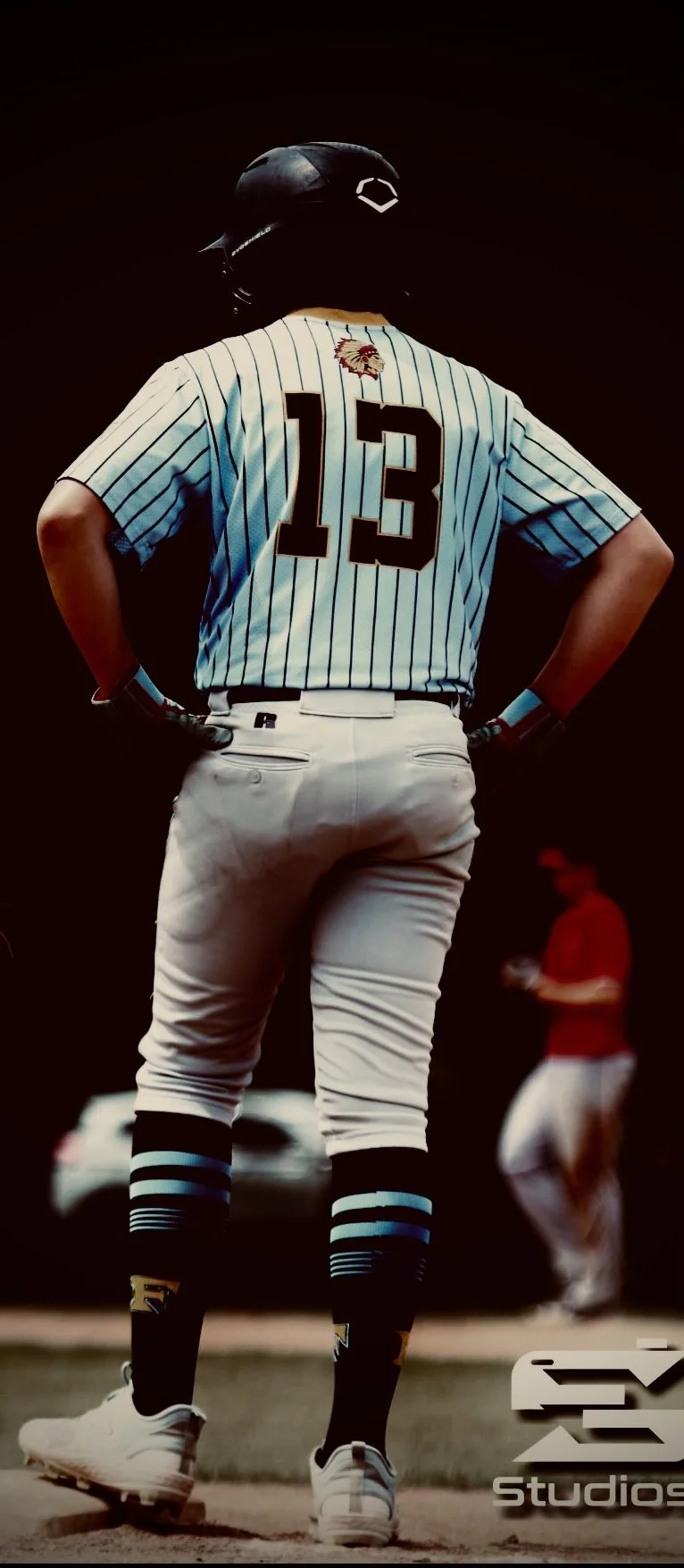 Baseball player wearing a jersey with the number 13, standing on the field with hands on hips, facing away, wearing a helmet and baseball cleats, with a person in a red shirt in the background.
