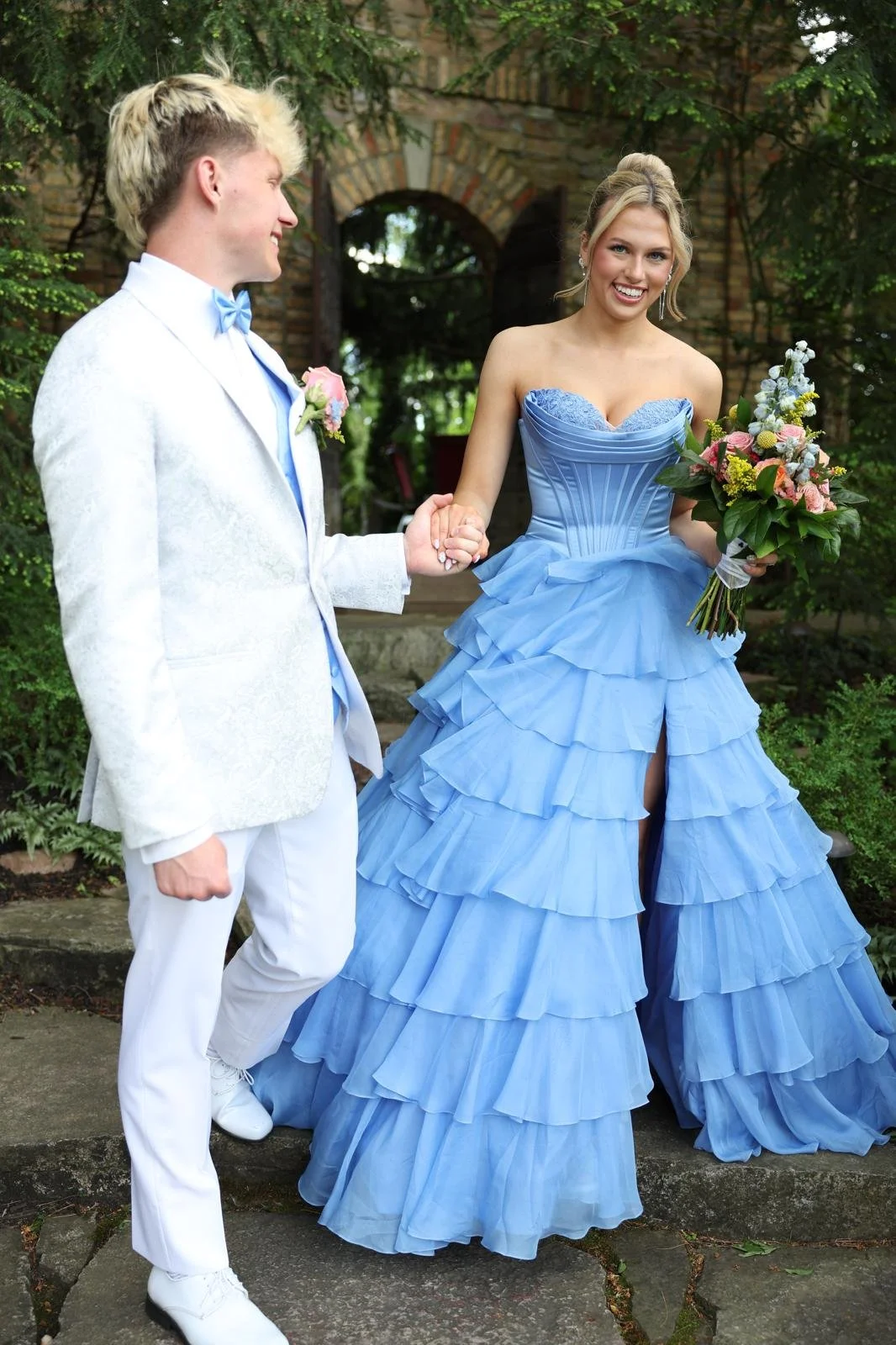 A bride and groom holding hands outdoors on a wedding day with greenery and an old brick building in the background. The bride is wearing a blue ruffled gown and holding a bouquet, smiling at the groom. The groom is dressed in a white suit with a blue bow tie and boutonniere.