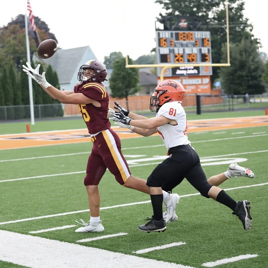 Football players in action on field, with one attempting to catch the ball as the other reaches for it, scoreboard in background.