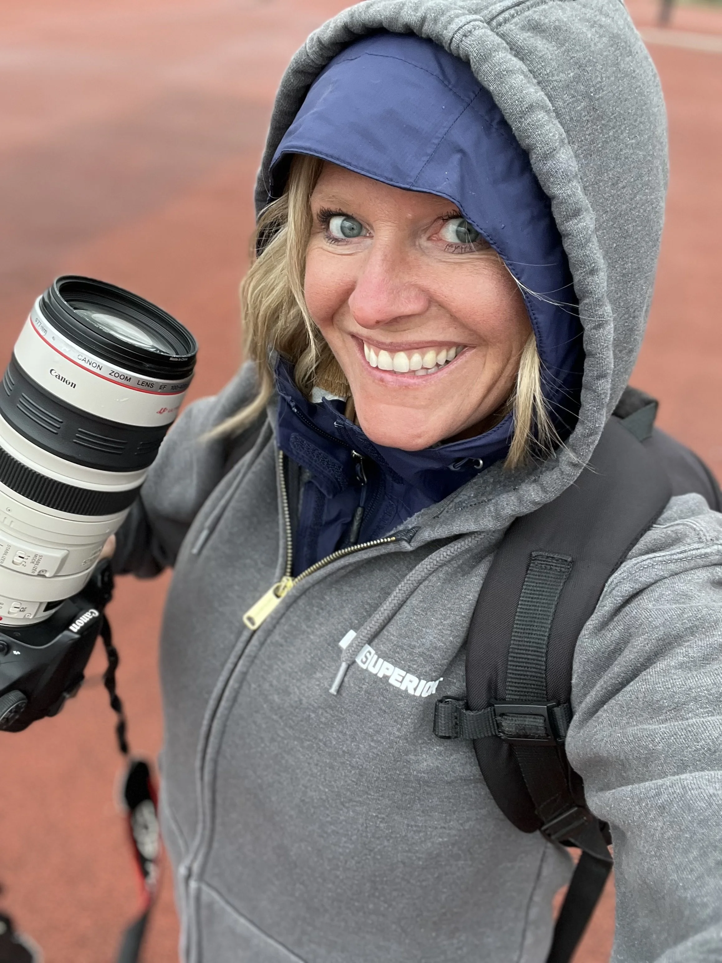 A woman with blue eyes and blonde hair taking a selfie outdoors on a reddish ground, wearing a gray hoodie with the hood up, a blue jacket under it, and carrying a black backpack, holding a Canon camera with a large lens.