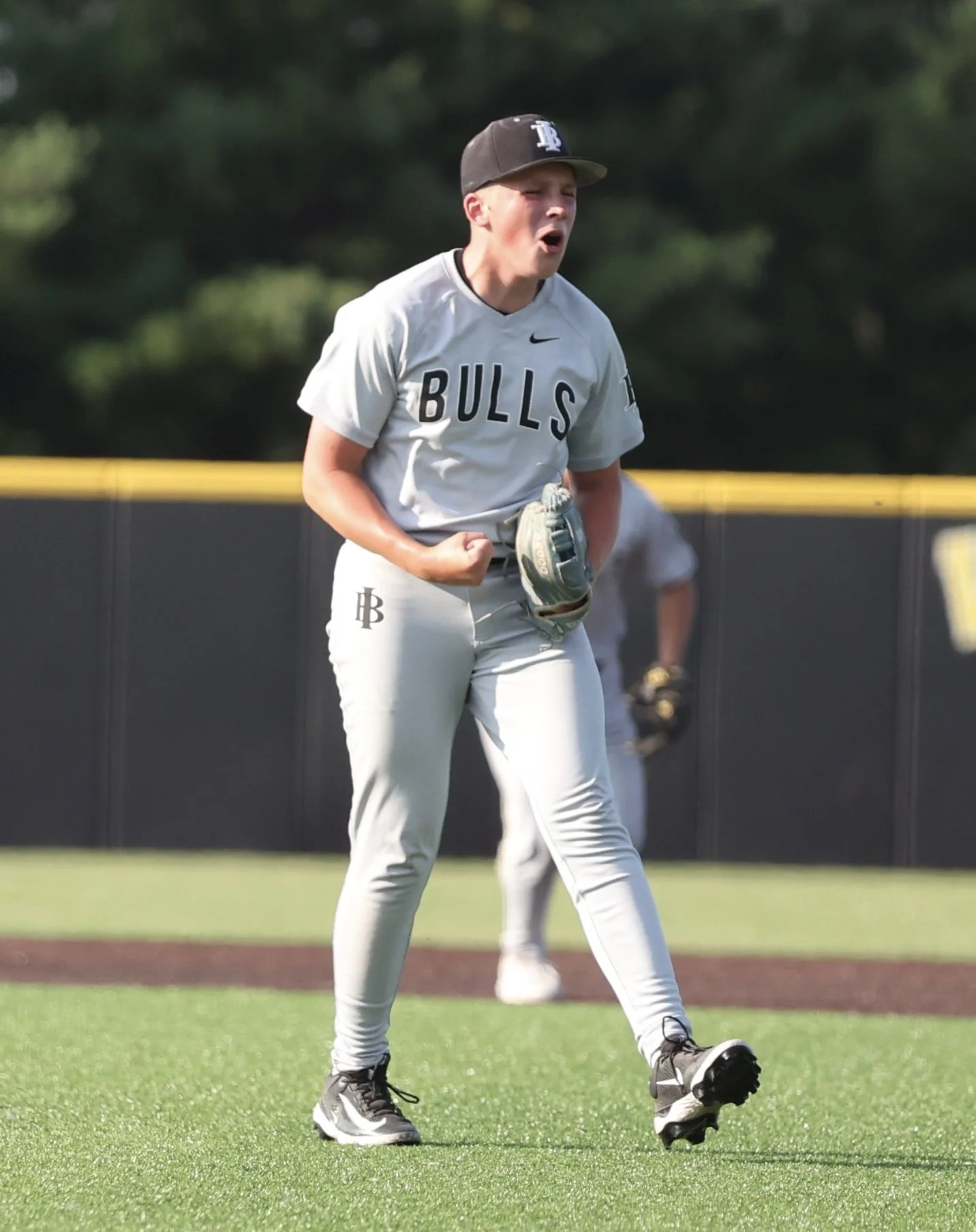 Young baseball player in gray uniform with 'BULLS' lettering, celebrating on the field with clenched fist, wearing a cap and glove, on a green grass field.